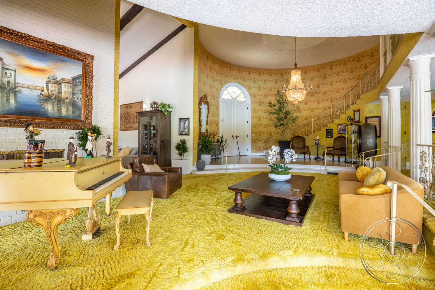 Euphoria - exposed brick walls, high vaulted ceiling, open living space, decorative potted plants, brown leather sofa, wooden accent table, gold-framed wall art, accent lighting fixtures, golden shag carpet flooring, round decorative chandelier, curved staircase with wood railing, large bay window, cream-colored armchairs, ornate gold mirror