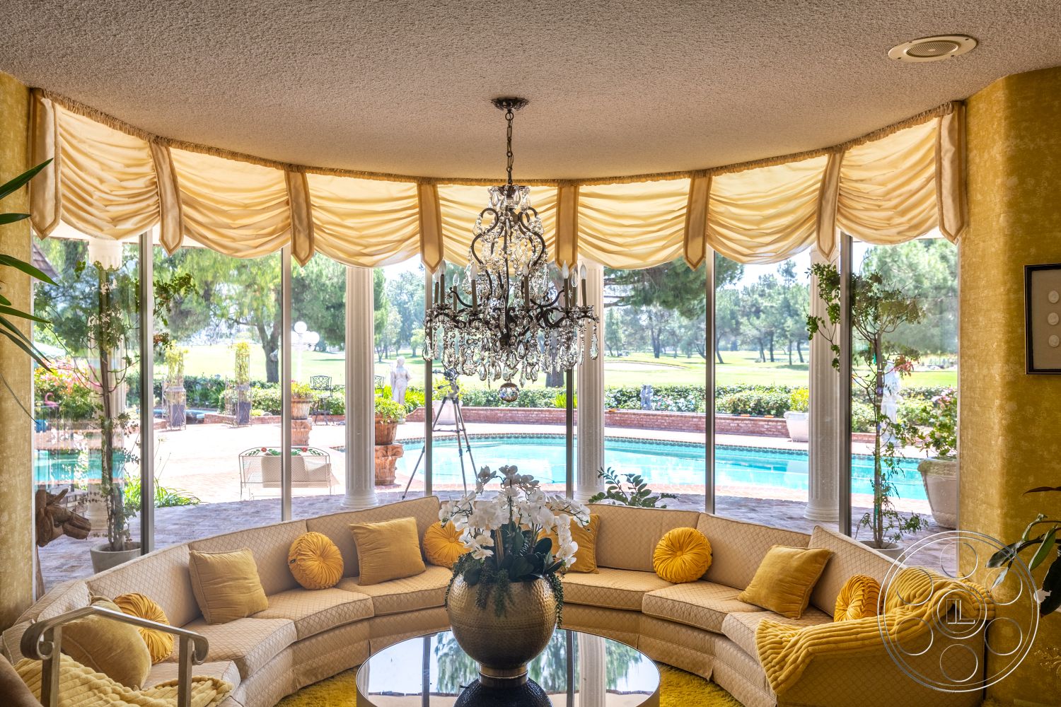 Euphoria - exterior, indoor plants, cozy seating area, floor-to-ceiling windows, textured ceiling, circular coffee table, swimming pool view, chandelier lighting fixture, marble patio flooring, rounded sectional sofa, yellow fabric drapery, golf course landscape