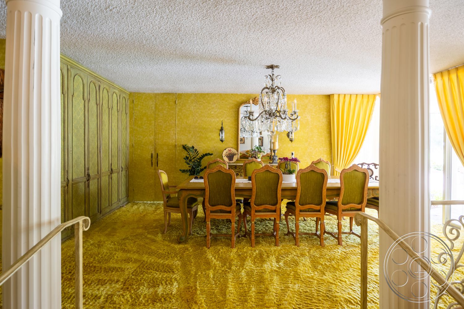 Euphoria - upholstered dining chairs, floor-to-ceiling windows, dark wood ceiling, walnut dining table, chandelier lighting fixture, built-in wooden cabinetry, decorative wall mirrors, ornate crown molding details, golden shag carpet flooring, yellow textured walls, curtains with yellow drapes, column structural supports