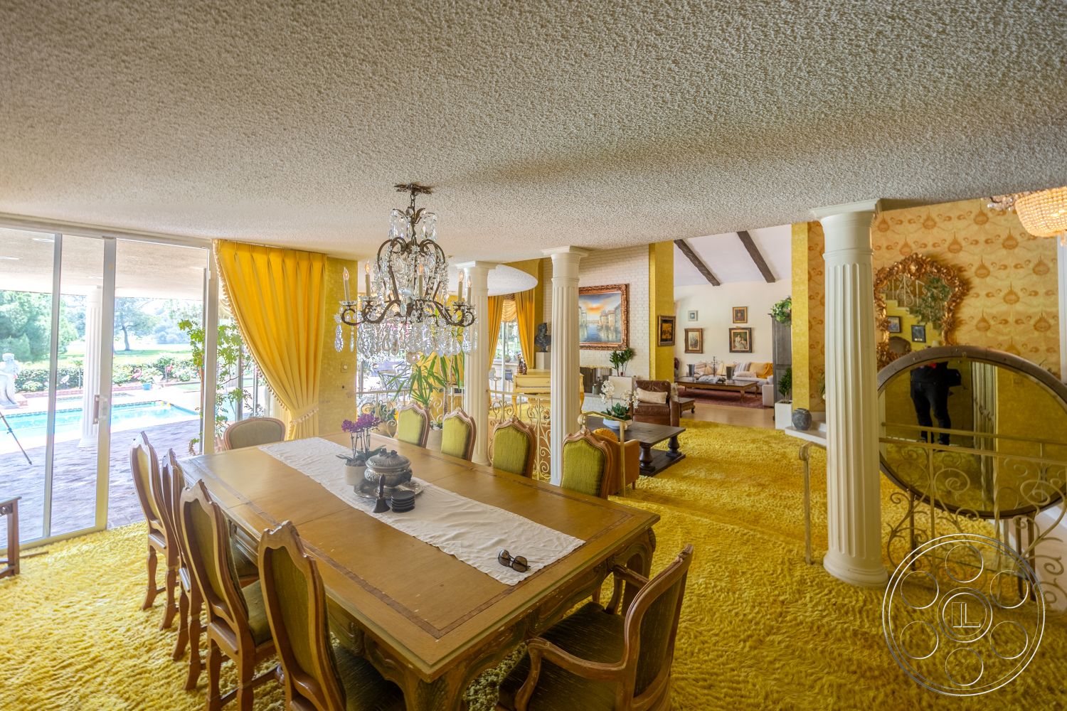 Euphoria - open floor plan, exposed beam ceiling, dining area, upholstered dining chairs, green plant accents, wood dining table, high ceiling, carpet flooring, colorful artwork, chandelier lighting, gold accent decor, living room view, large sliding glass doors, decorative wall paneling, textured ceiling finish, yellow drapery, view of pool area, column architectural details