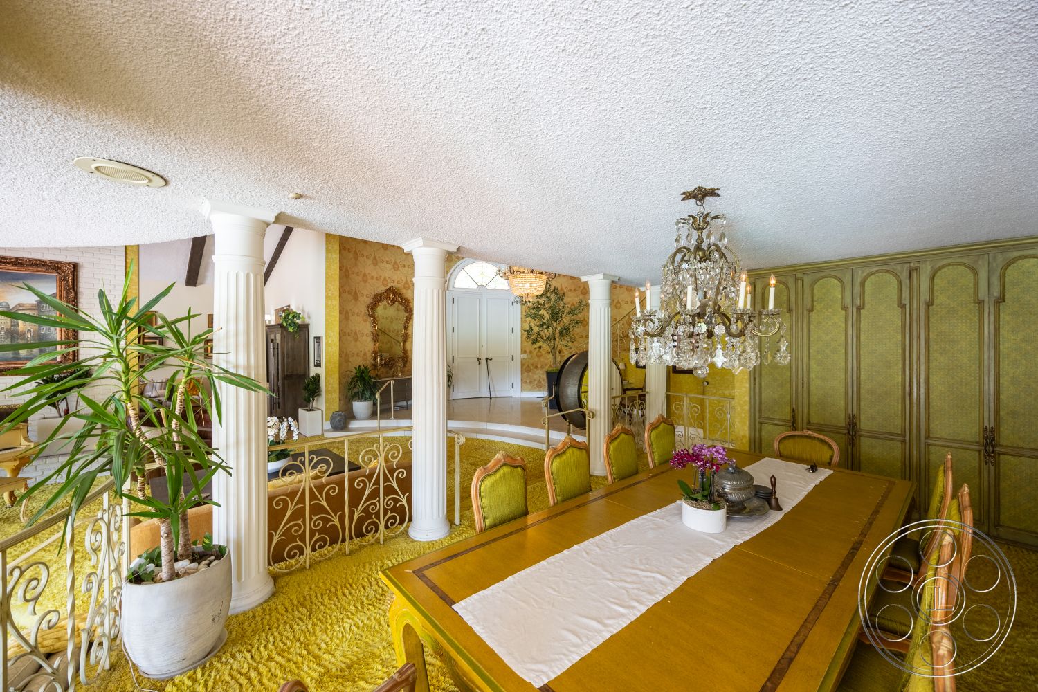 Euphoria - carpet flooring, textured plaster ceiling, decorative columns, interior dining area, open floor plan layout, chandelier with crystal accents, hardwood dining table, plant in ceramic pot, upholstered green dining chairs, gold and green wallpaper, built-in cabinetry with detailed trim, vintage-style decor, ornate mirror wall accent