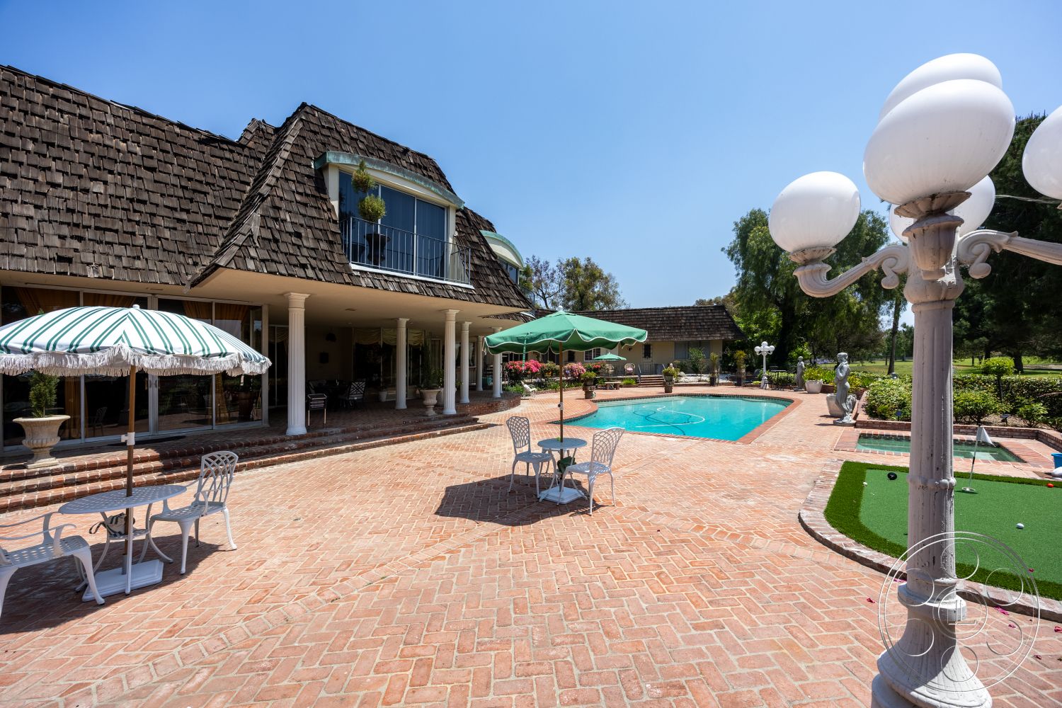Euphoria - exterior, rectangular pool, stone planter, golf putting green, landscaped garden setting, brick patio flooring, wood shingle roof, white column supports, green and white striped umbrellas, white metal patio furniture, decorative outdoor lamp post, poolside paving stones, flowering plants around pool area