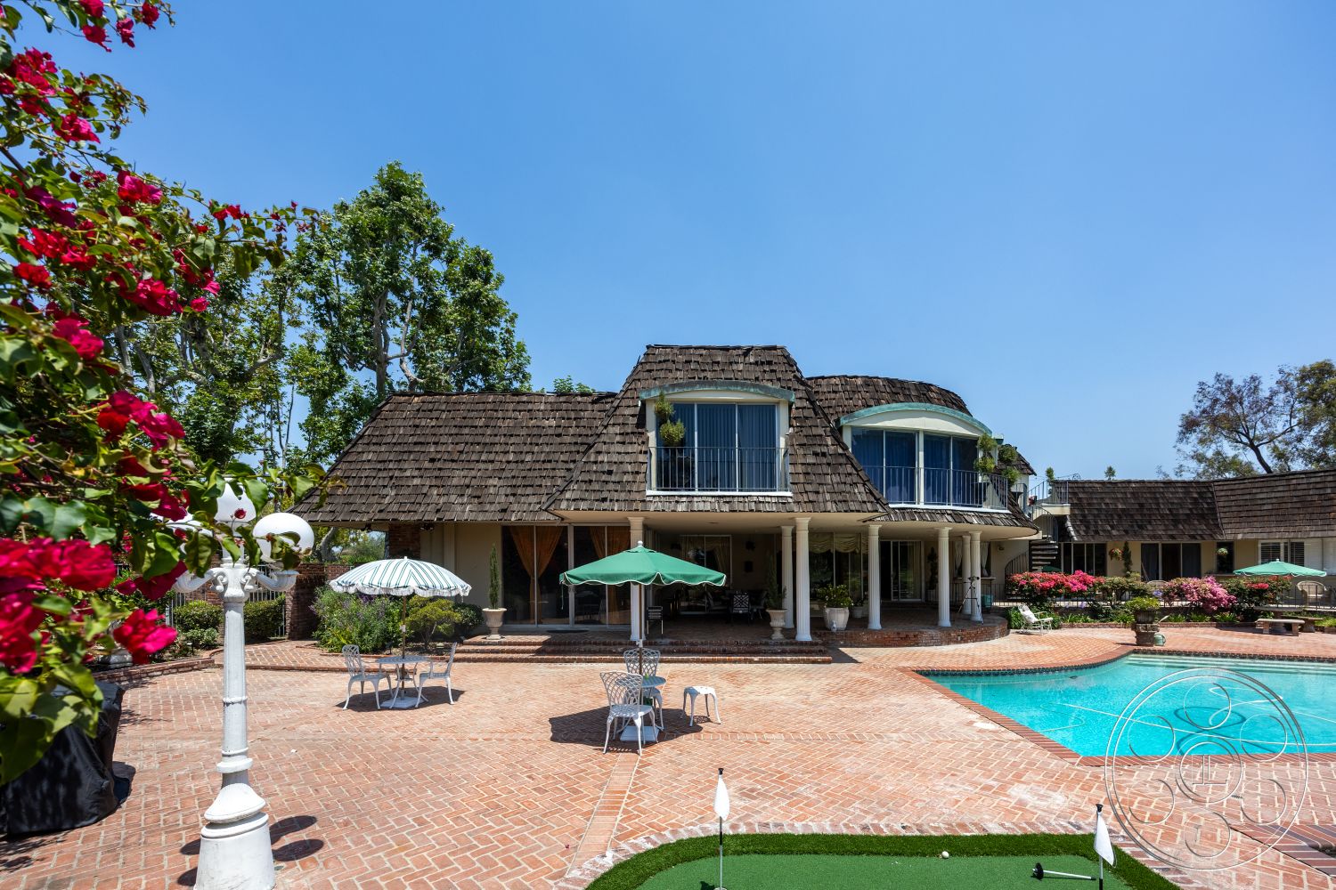 Euphoria - exterior, landscaped yard, rectangular swimming pool, white columns, flowering shrubs, wood deck, stone walkway, arched windows, manicured garden, modern farmhouse style, poolside seating area, shingle roof, sunny outdoor setting, two-story home, curved pool edge, brick patio flooring, outdoor dining furniture, green umbrellas