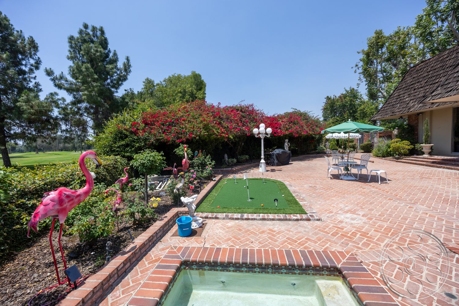 Euphoria - exterior, sitting area, outdoor lighting fixtures, landscape garden, brick patio flooring, lush tropical vegetation, manicured garden setting, artificial grass putting green, decorative pink flamingo lawn ornaments, lush greenery with flowering shrubs, table and chairs with an umbrella, traditional brick border, concrete spa with tiled edge, shingled roof on adjacent structure