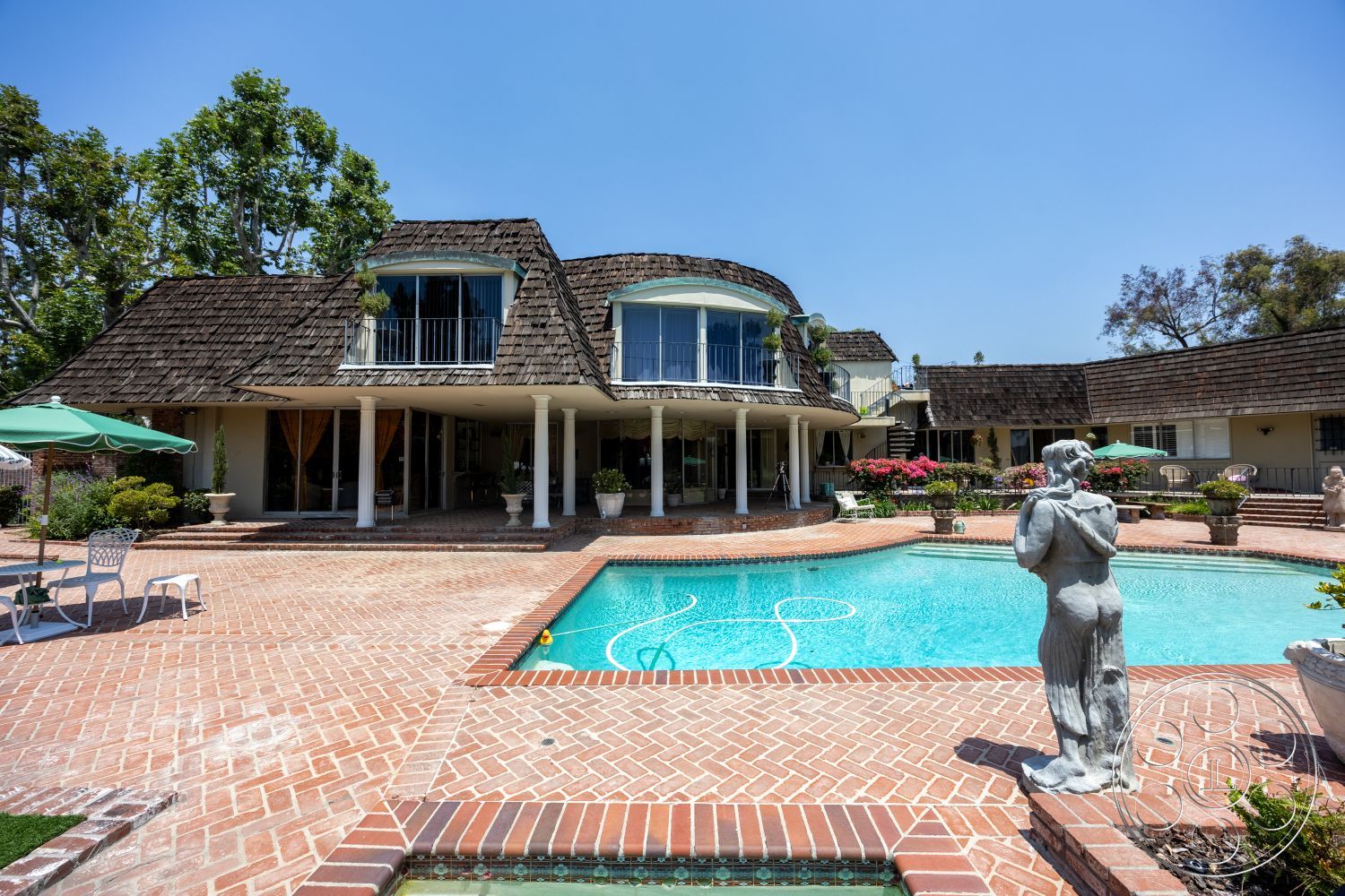 Euphoria - exterior, sunny outdoor space, two-story home, ornamental statue, oval swimming pool, stucco exterior walls, curved balcony railing, wood shingle roof, landscape garden setting, herringbone brick patio flooring, planter pots with flowers, wooden deck chairs, green patio umbrella, vaulted roof design, freestanding columns, pavers around pool area