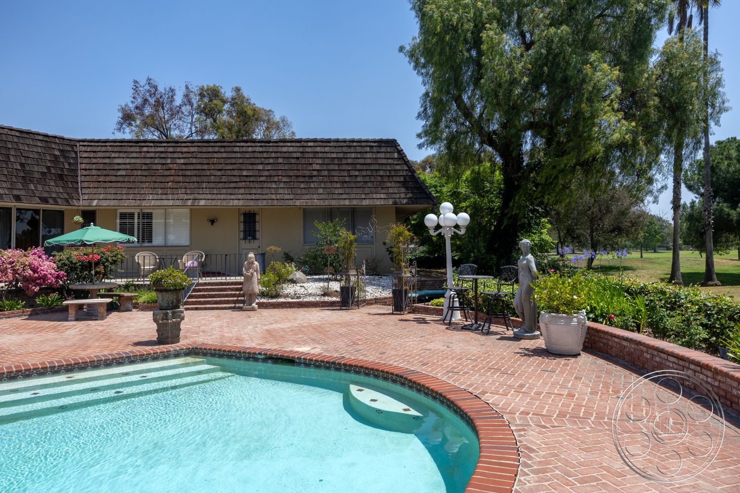 Euphoria - exterior, landscaped garden, residential backyard, metal patio furniture, oval swimming pool, lush greenery setting, concrete pool border, brick patio flooring, terracotta flower pots, wood shingle roof, green patio umbrella, sculptural garden accents