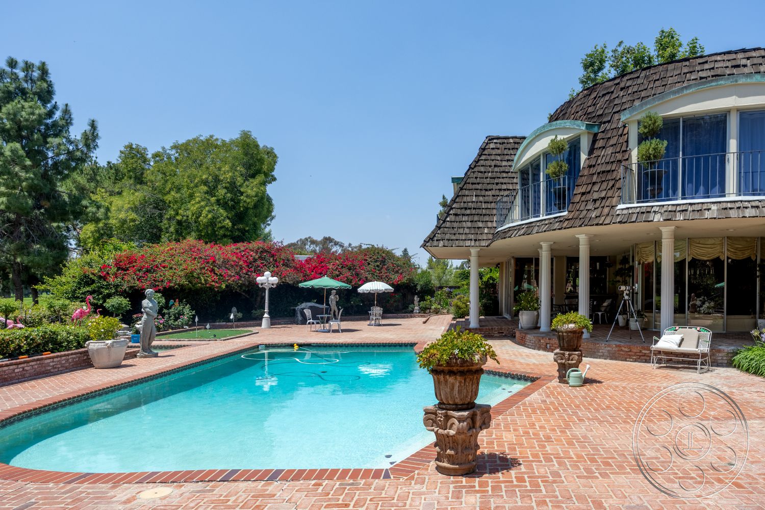 Euphoria - exterior, large glass windows, outdoor dining area, poolside lounge chairs, cobblestone path, stone planter, brick planters, rectangle swimming pool, natural greenery backdrop, brick patio flooring, half-timbered roof with shingles, lush floral landscaping, tall garden statues, circular umbrella shade, arched porch
