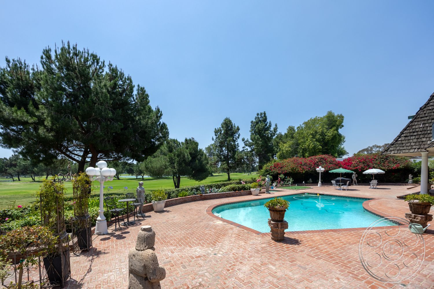 Euphoria - exterior, outdoor seating area, rectangular swimming pool, planters with greenery, tall pine trees, brick patio flooring, sunny backyard setting, golf course view, wrought iron patio furniture, lush landscaping with flowering shrubs, ornamental stone sculptures, freestanding outdoor lanterns, umbrella-shaded tables