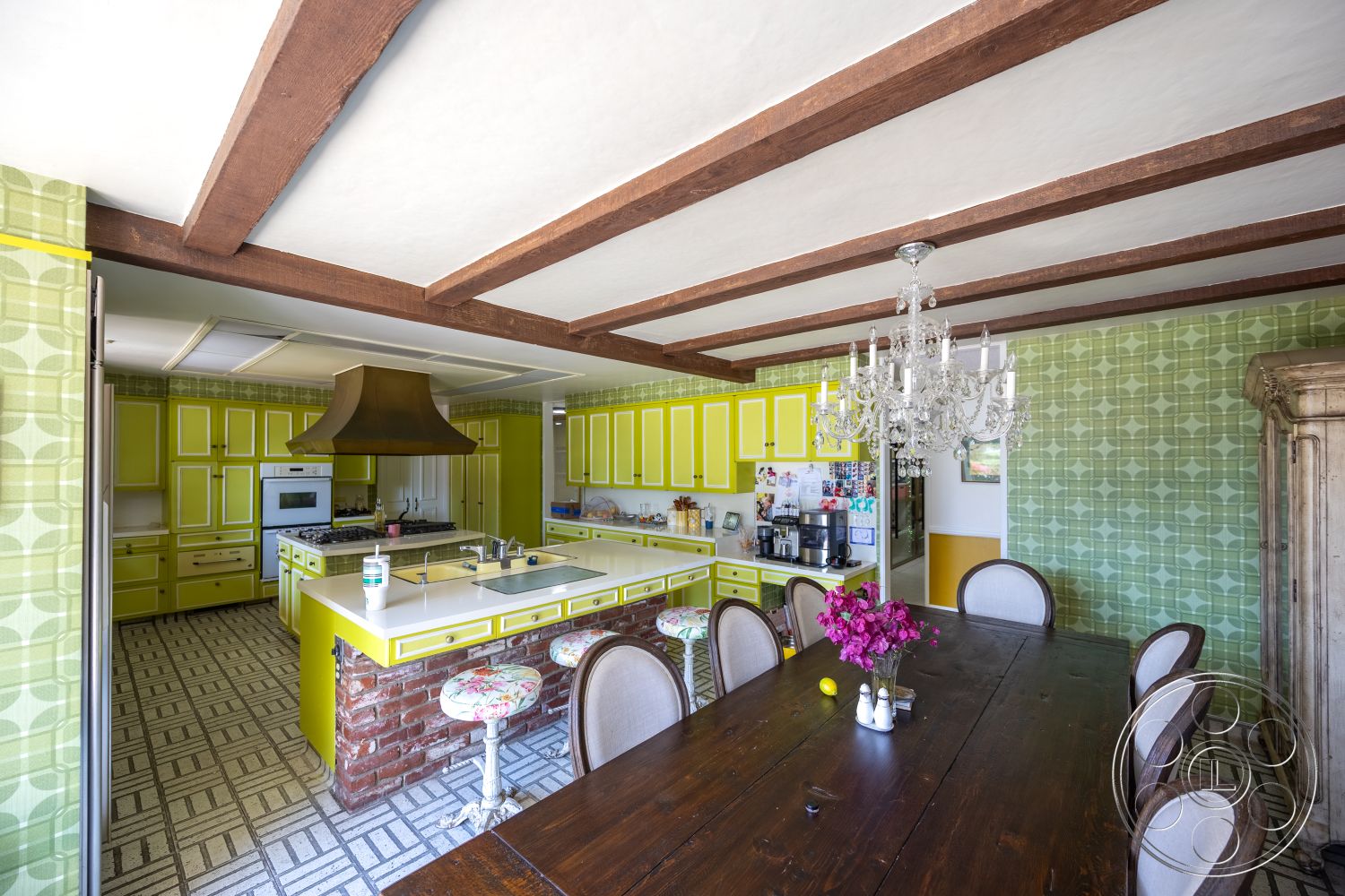 Euphoria - stainless steel appliances, upholstered dining chairs, wood dining table, ceramic tile flooring, textured plaster walls, chandelier light fixture, wood beam ceiling, quartz kitchen countertop, yellow kitchen cabinets, brick kitchen island, green patterned wallpaper