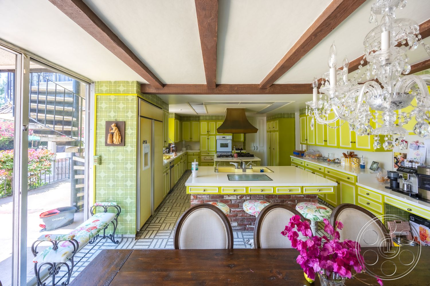Euphoria - yellow cabinetry, dark wood dining table, glass sliding door, wood beam ceiling, chandelier lighting fixture, concrete floor tiles, brick kitchen island, green geometric backsplash, stainless steel kitchen appliance, hanging copper range hood, floral upholstered stool, open layout kitchen design
