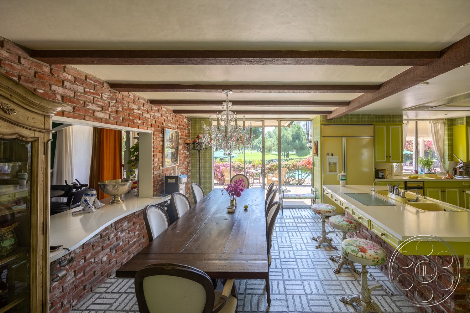 Euphoria - large glass sliding doors, open floor plan, dark wood dining table, chef's kitchen, natural light-filled space, wood beam ceiling, exposed brick accent wall, tile kitchen floors, vintage-style bar stools with floral upholstery, modern kitchen cabinets with yellow finish, interior chandelier, bright and airy dining area