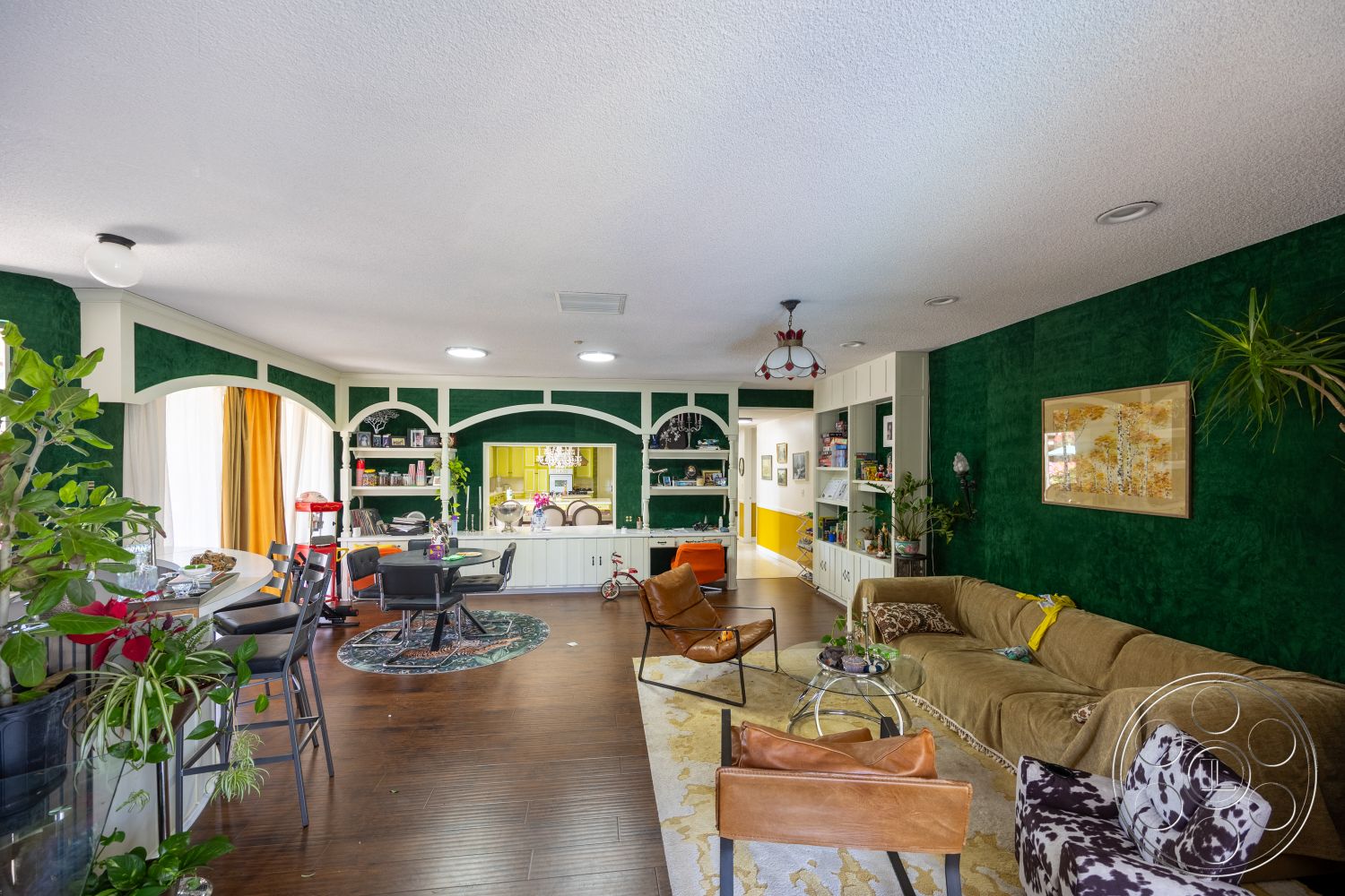 Euphoria - decorative area rug, indoor plants, glass coffee table, hanging light fixture, white built-in shelving, plush brown sofa, hardwood living room floors, yellow accent wall, green textured wall treatment, arched wall details, wrought iron bar stools, large windows with curtains