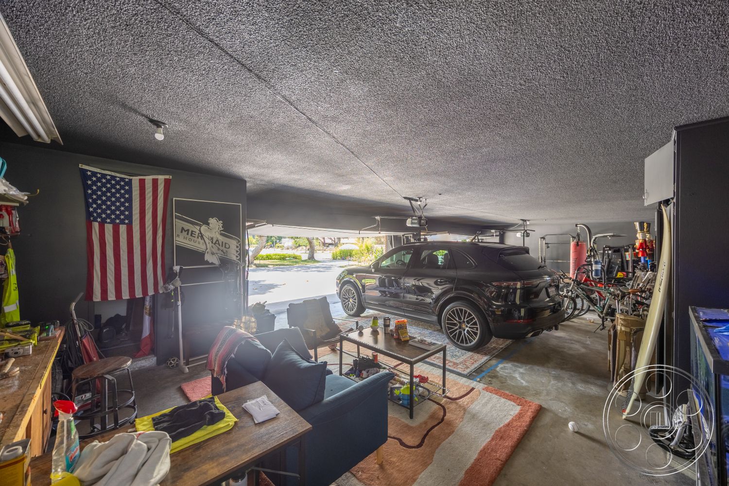 Euphoria - exposed ceiling, painted walls, overhead lighting fixtures, area rug with geometric pattern, reclining chairs, concrete garage floor, interior garage space, black SUV vehicle, wood workbench, pulley system on ceiling, American flag wall decoration, stylized mermaid sign, shelving for tools, miscellaneous storage items