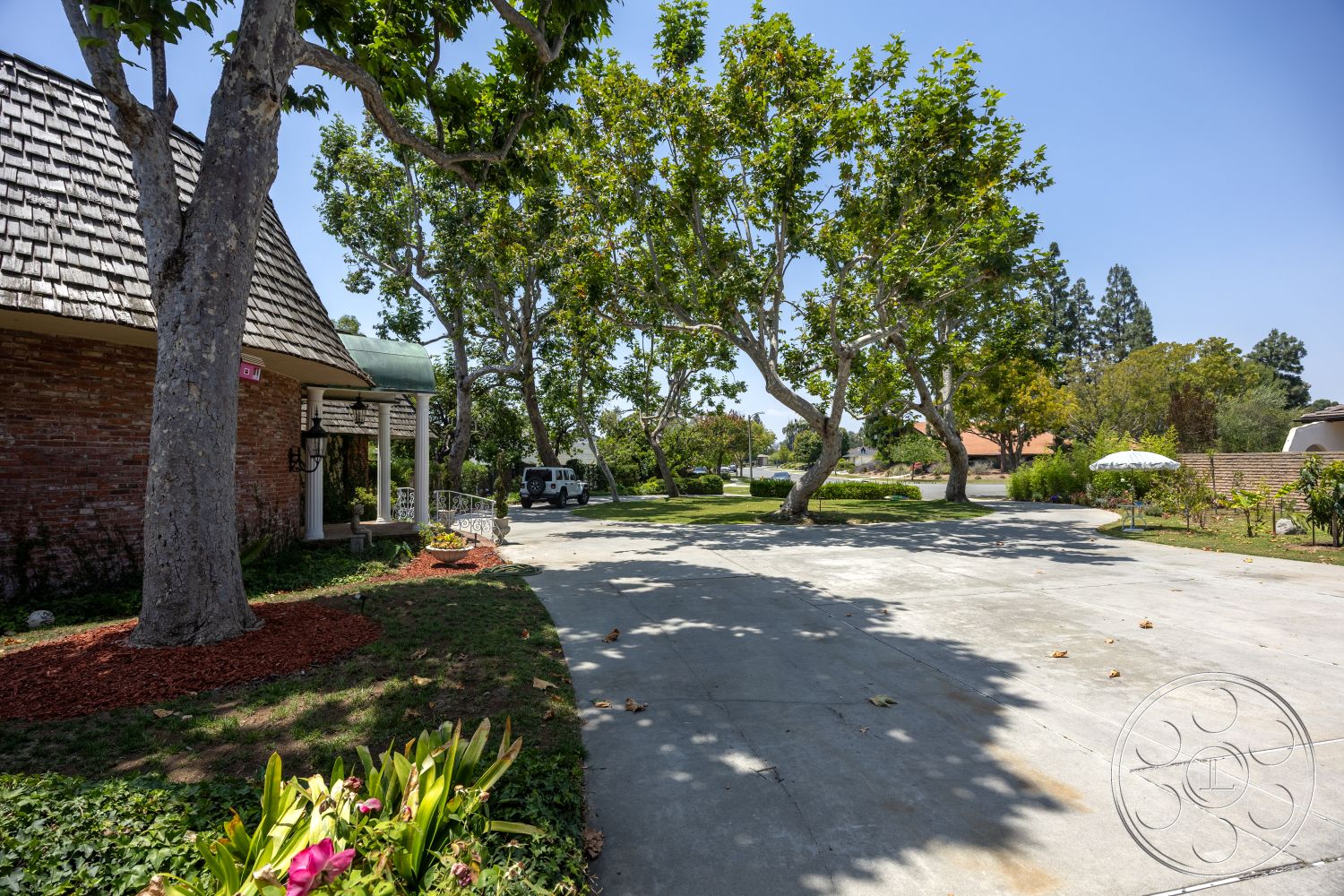 Euphoria - exterior, mature trees, stone pathway, concrete driveway, shingle roof, landscaped garden area, brick wall facade, flower bed with colorful flowers, decorative garden mulch, sunny suburban setting