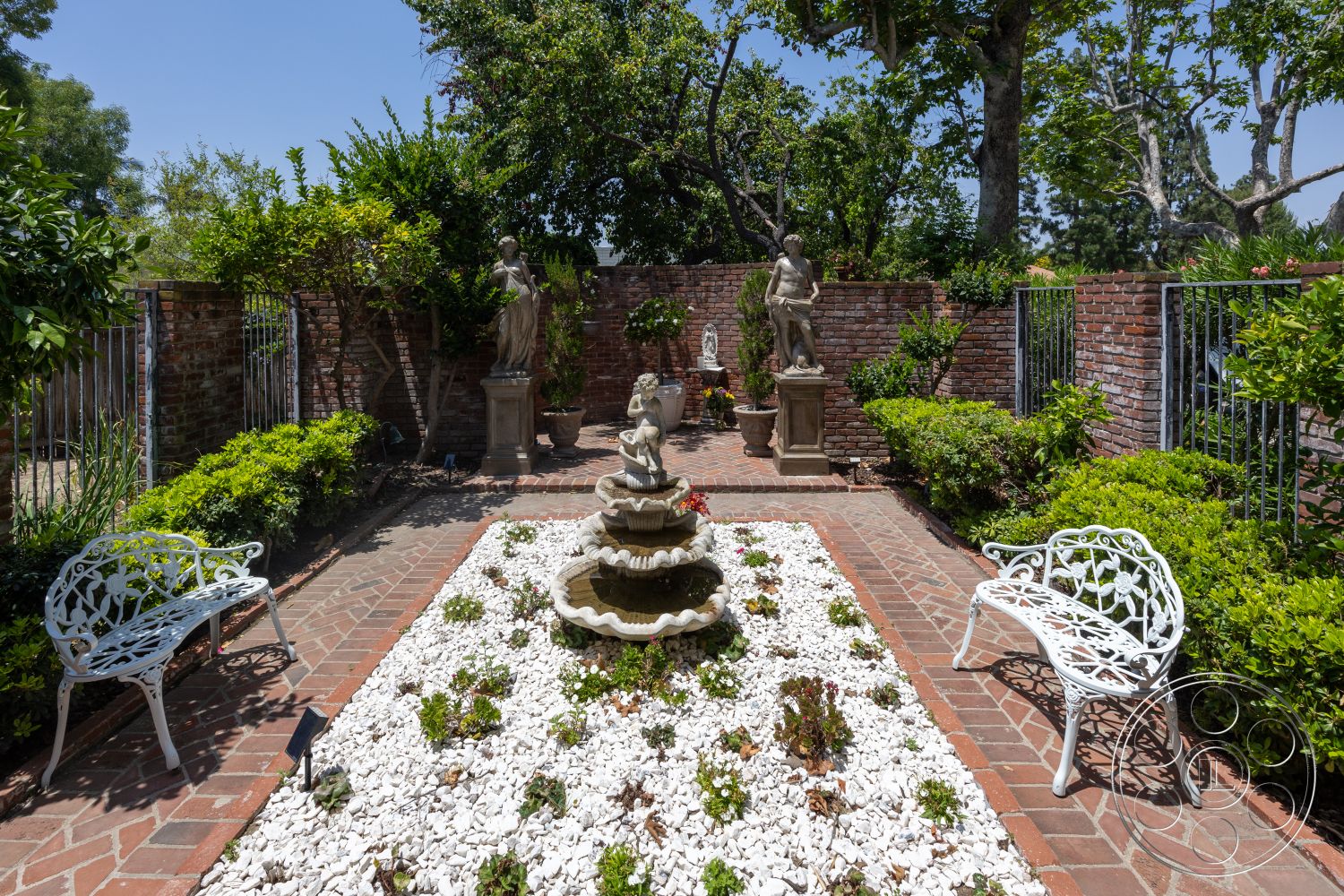 Euphoria - exterior, lush greenery, green hedges, trees providing shade, brick garden walls, red brick pavers patio, white decorative gravel ground cover, cast stone fountain, ornamental statues, white wrought iron garden benches