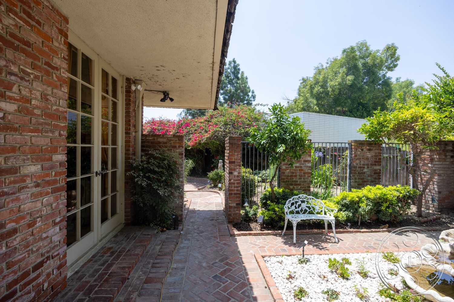 Euphoria - exterior, landscaped garden, patio area, brick pathway, paved walkway, green shrubbery, flowering plants, textured plaster walls, exposed concrete ceiling, French doors, red brick walls, iron gate fencing, white ornamental bench, terracotta flower bed