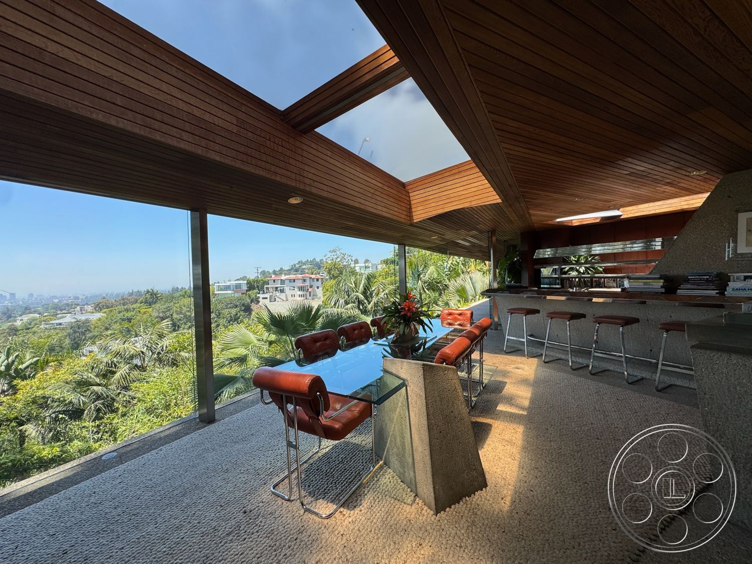 Goldstein House - open concept layout, surrounding greenery, floor-to-ceiling windows, glass dining table, woven area rug, mid-century modern style, exposed wood ceiling beams, leather dining chairs, granite kitchen countertops, concrete accent wall, sloped skylight, panoramic garden view