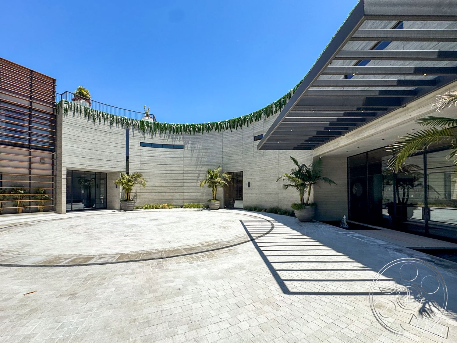 Star Villa - exterior, large outdoor space, sliding glass windows, modern architectural design, glass entrance doors, curved concrete walls, concrete circular driveway, bright sunny environment, wood slatted facade, landscape planters with palm trees, stone paver flooring, overhanging metal roof
