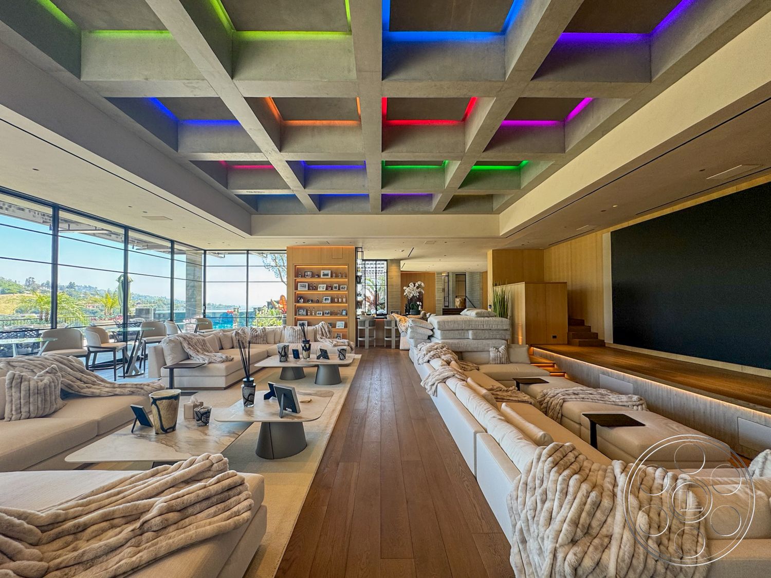 Star Villa - neutral color palette, contemporary decor, open floor plan, hardwood living room floors, decorative indoor plants, floor-to-ceiling glass windows, modern coffee tables, city skyline view through windows, built-in wooden shelving unit, coffered concrete ceiling with colorful LED lighting, white sectional sofas, textured throw blankets on sofas, multi-level wooden platform