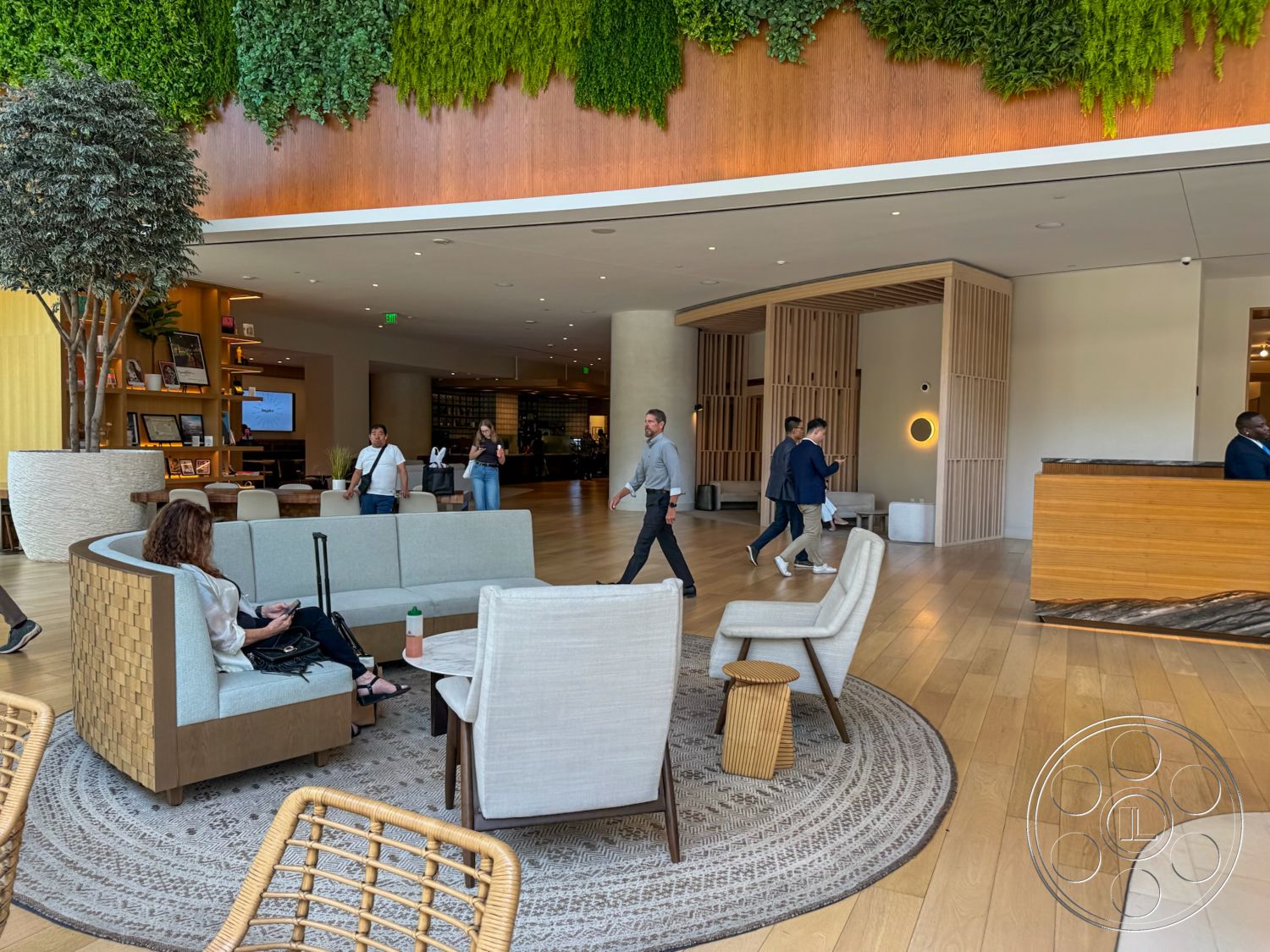 Office 123 - open floor plan, light wood flooring, modern coffee table, floor-to-ceiling windows, contemporary seating arrangement, wood panel wall treatment, built-in wooden shelving unit, sofa with curved shape and tan upholstery, decorative round area rug, hanging indoor plants, reception desk with natural wood finish