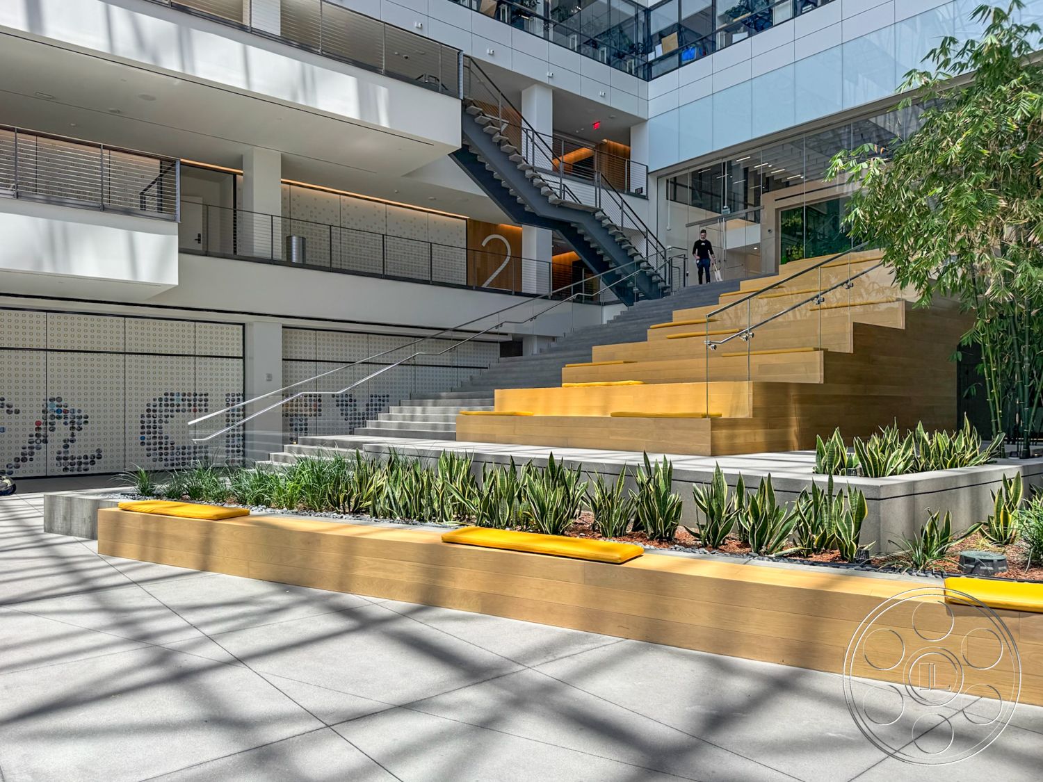 The Atrium - indoor greenery, glass balcony railing, open atrium design, concrete floor tiles, exposed steel structural elements, wooden step seating, stainless steel staircase, landscaped planter boxes, yellow accent seating, multi-level seating area