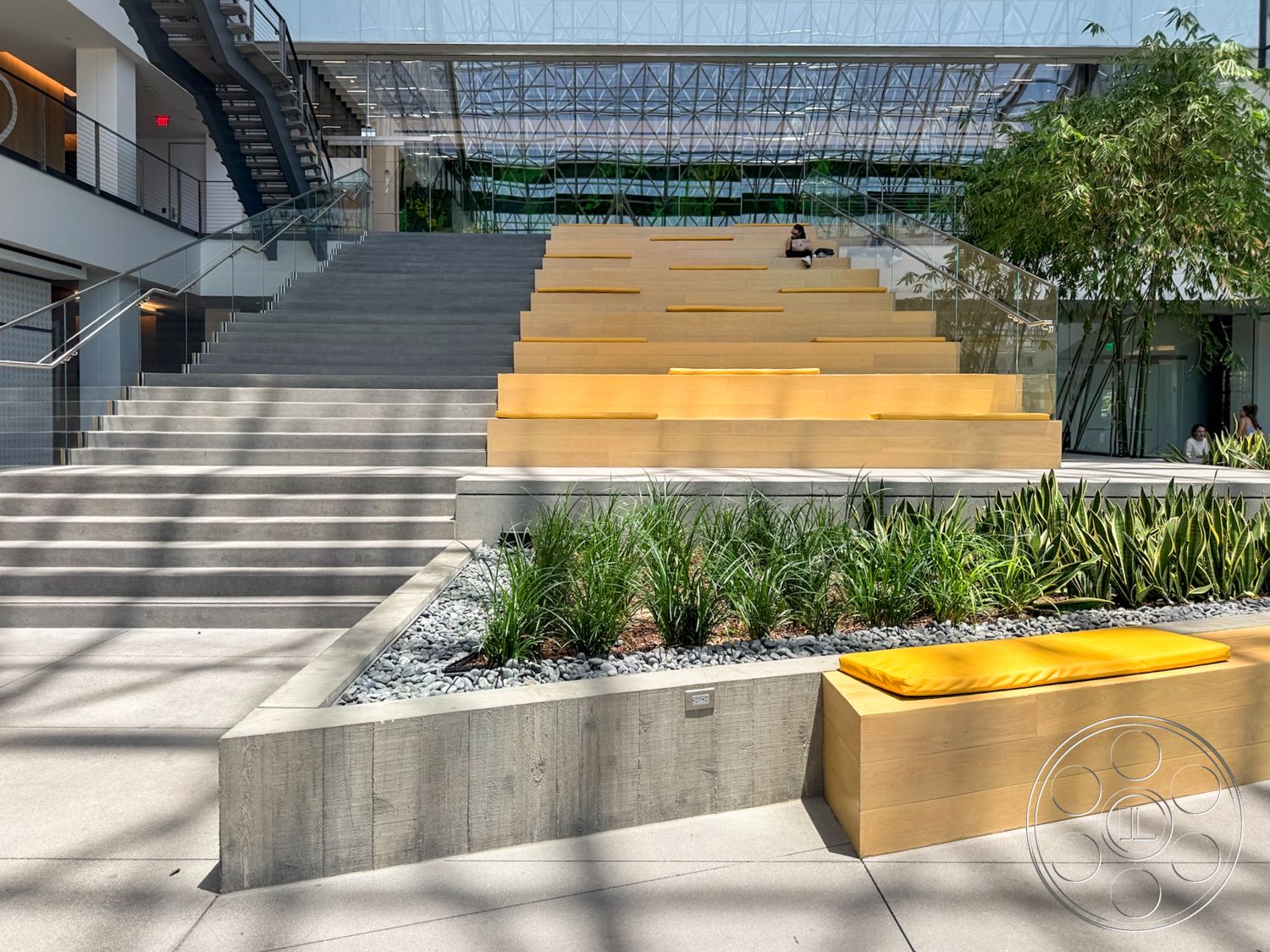 The Atrium - exterior, glass railing, concrete steps, textured concrete flooring, modern architectural style, steel staircase, concrete planter box, wood seating with yellow cushions, green landscaping with low-growing plants, linear geometric design, open-air atrium design, natural light through glass roof, horizontal seating arrangement
