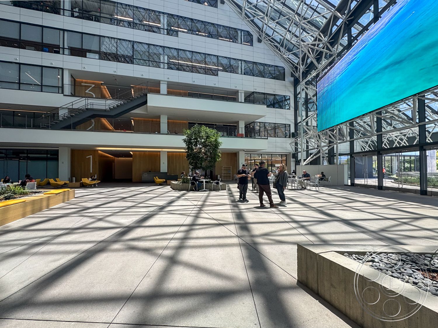 The Atrium - exterior, open floor plan, concrete flooring, modern architectural style, light-filled space, glass curtain wall, large digital display screen, two-story atrium, steel structural framework, landscaped planter boxes, indoor tree presence, yellow upholstered seating, decorative stone aggregate landscaping, interior gathering space