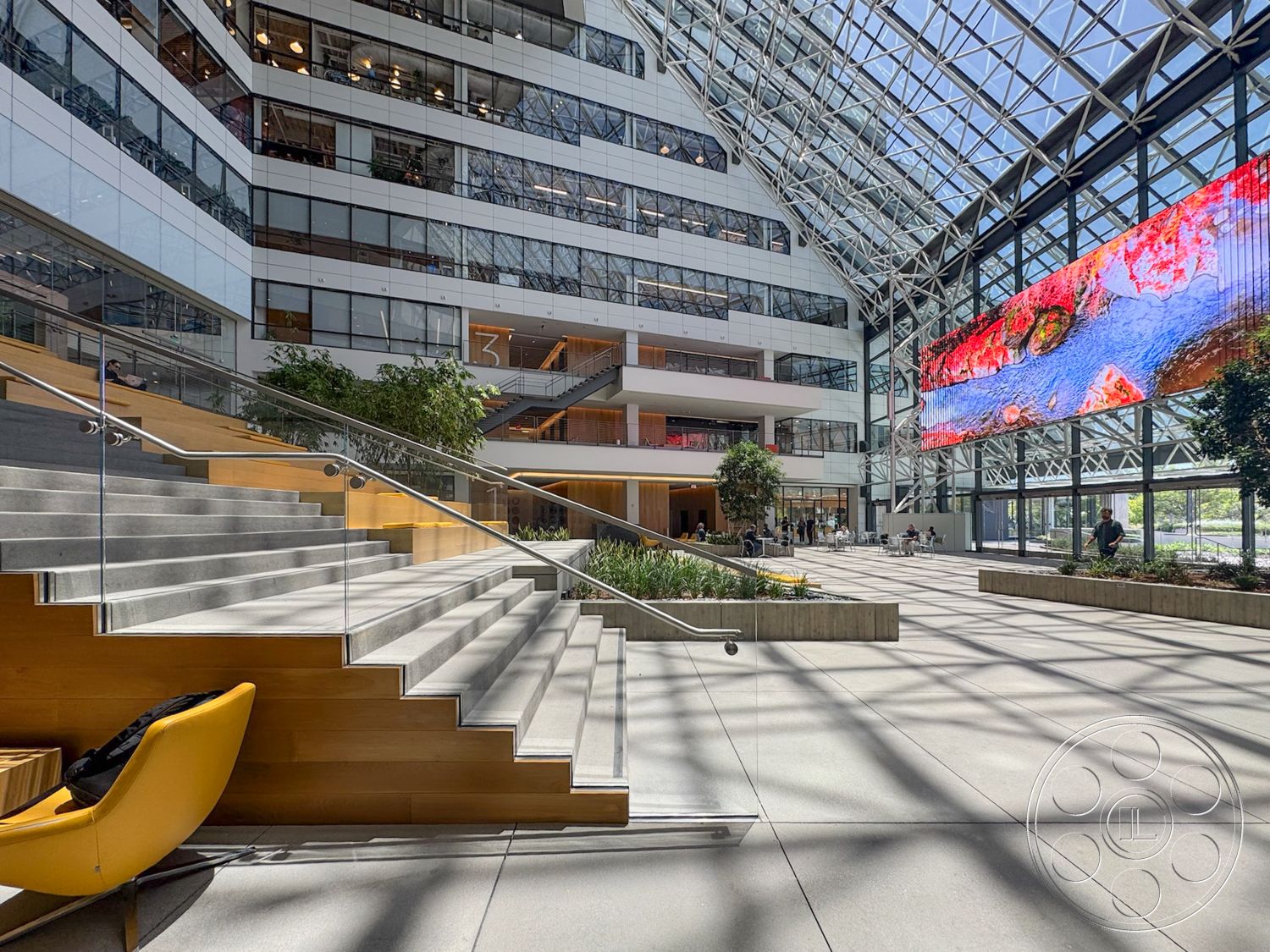 The Atrium - glass railings, hanging planters, bright natural lighting, indoor greenery, steel frame structure, exposed steel beams, wooden stair treads, open atrium space, modern office environment, digital display screen, paved concrete floor, concrete staircase, glass atrium roof, multilevel layout