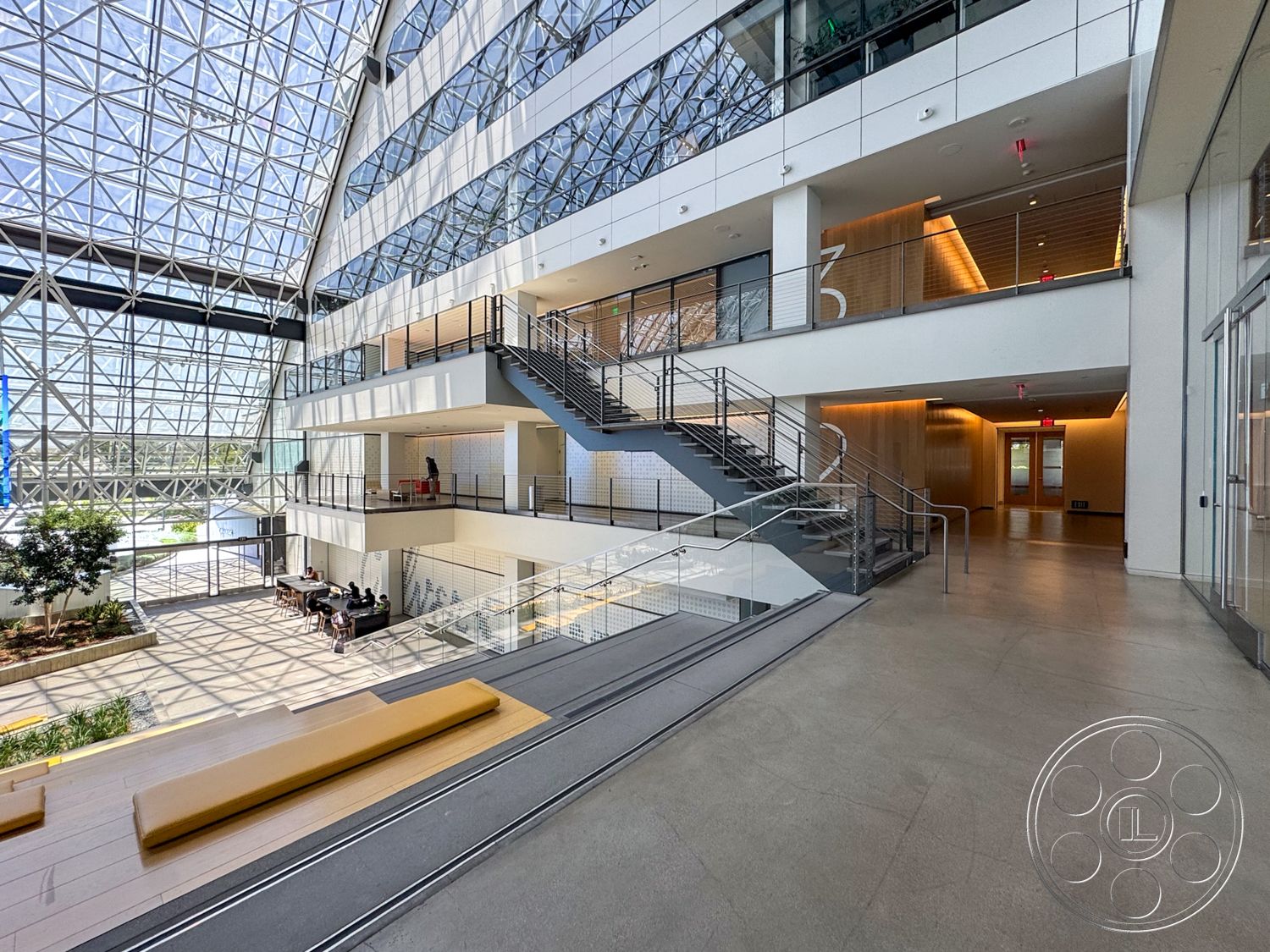 The Atrium - natural light, open floor plan, concrete flooring, modern architectural style, white painted walls, steel stair railings, glass atrium ceiling, exposed structural framework, wood seating platform, landscaped indoor area