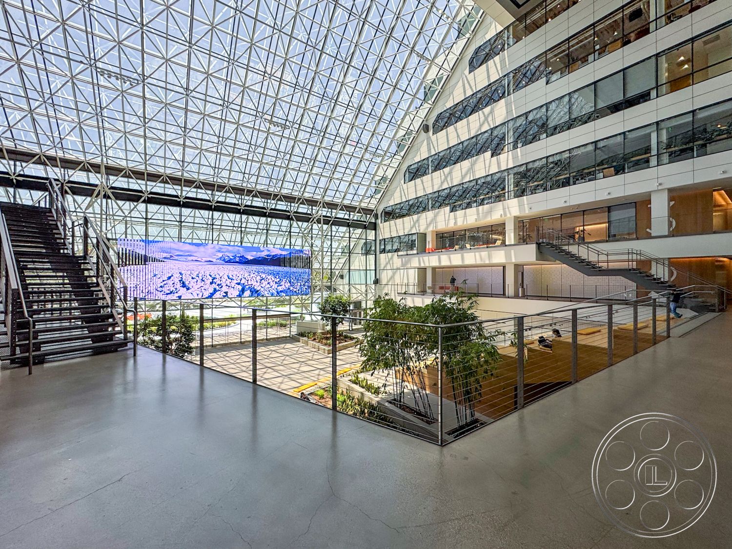 The Atrium - concrete floor, floor-to-ceiling windows, modern architectural style, metal railing details, large digital display screen, glass atrium ceiling, open courtyard area, steel framed staircase, landscaped indoor plants