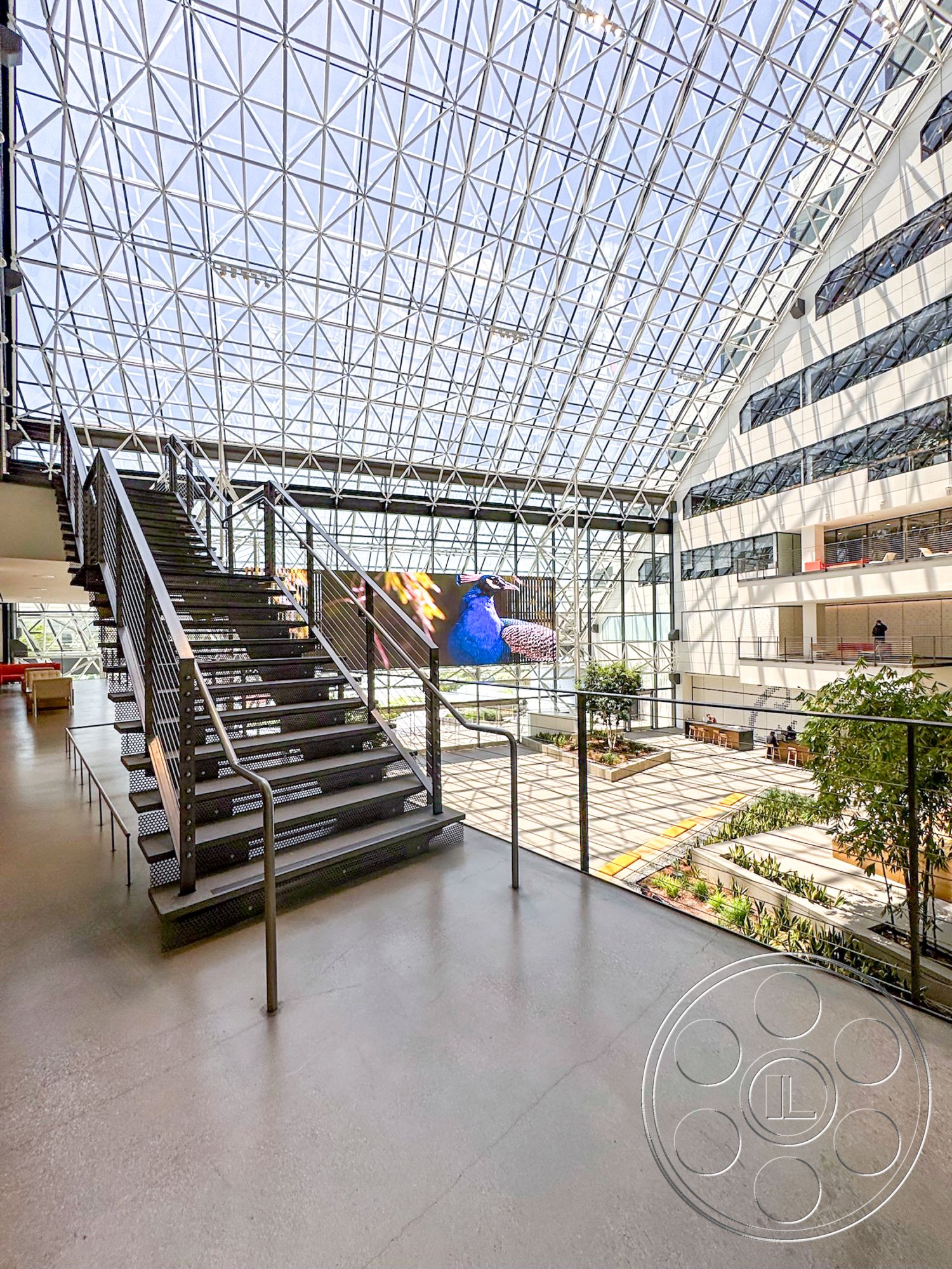 The Atrium - concrete flooring, large glass windows, art installation, modern architectural style, exposed framework, textured wall details, natural light access, metal stair railing, glass atrium roof, steel open staircase, landscaped indoor courtyard