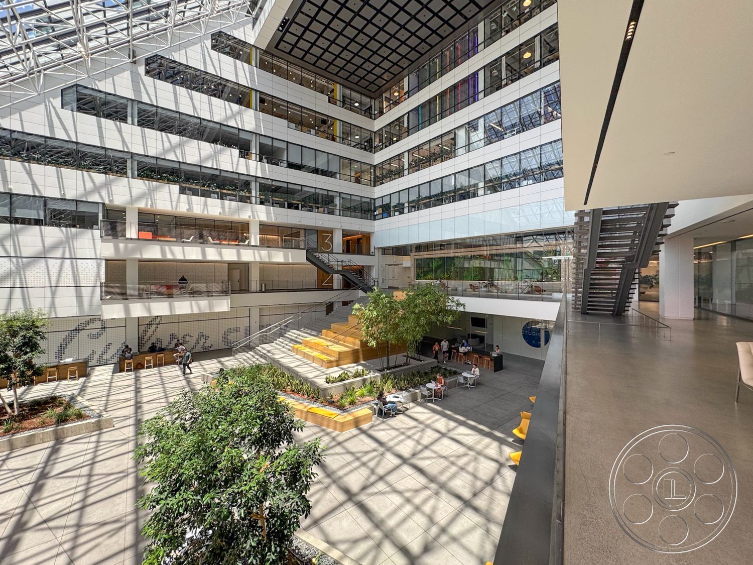 The Atrium - concrete flooring, minimalist design elements, floor-to-ceiling windows, planted greenery, modern architectural style, steel staircase, brightly lit interior, exterior courtyard view, glass atrium roof, wooden seating benches, open-air office layout, multiple levels of open space, industrial structural details