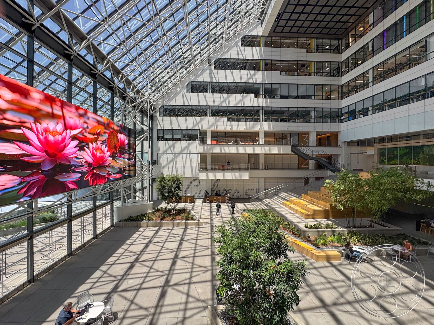 The Atrium - natural light, concrete flooring, textured concrete walls, floor-to-ceiling windows, industrial design elements, modern architectural style, stainless steel railings, exterior glass facade, open atrium space, landscaped planter beds, steel structural framework, large digital display wall, indoor garden area, wooden seating steps, multi-level atrium