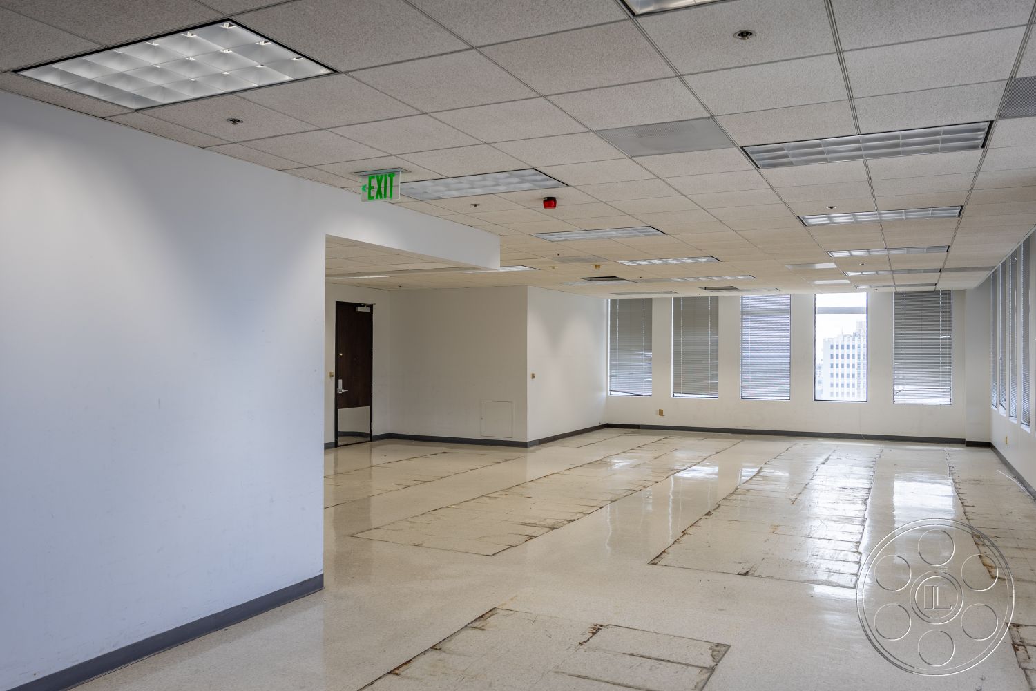 Rooftop 53 - large windows, horizontal blinds, white painted walls, drop ceiling tiles, fluorescent ceiling lights, sheet vinyl flooring, exposed concrete floor patches, fire exit sign