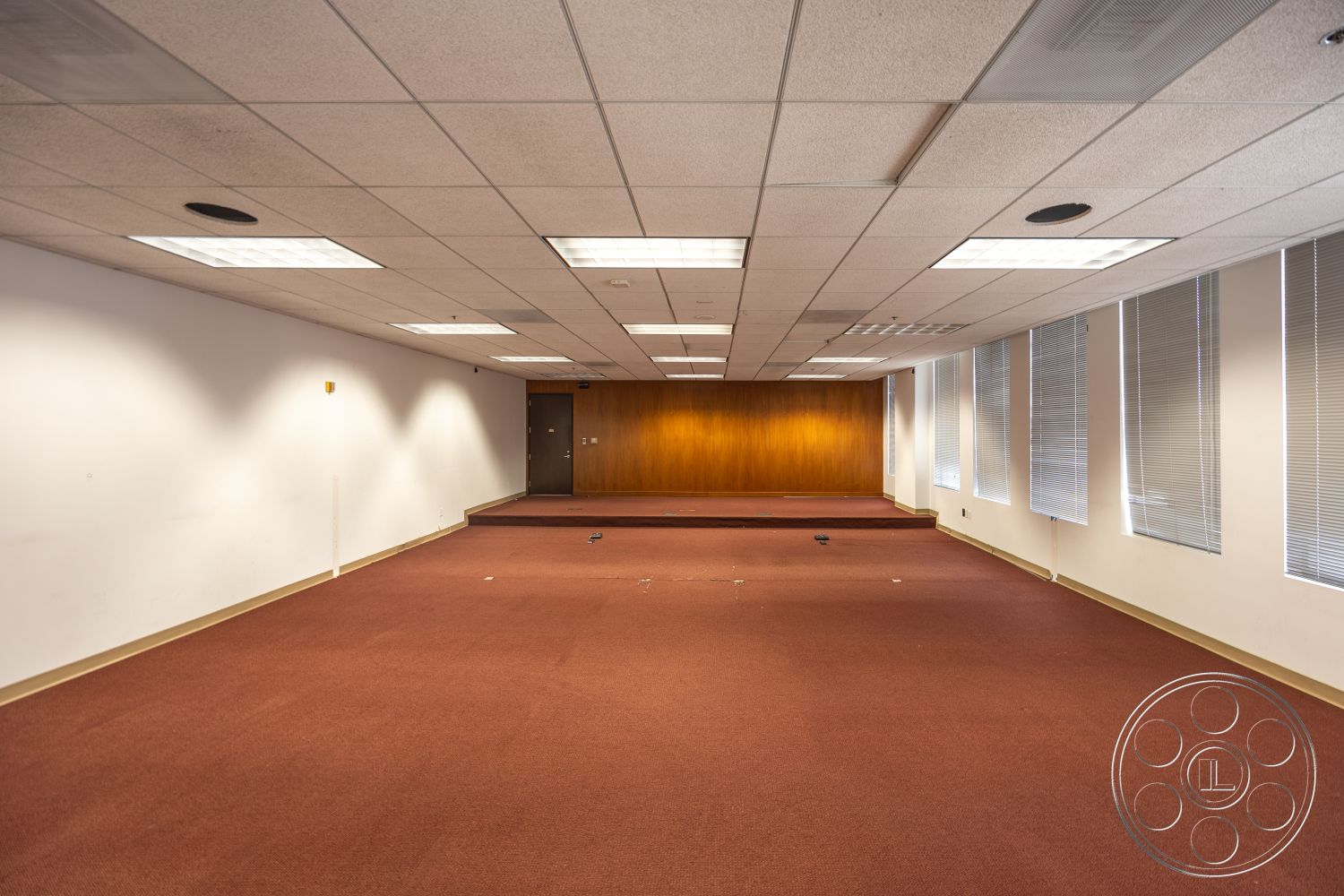 Rooftop 53 - recessed lighting, carpet flooring, neutral painted walls, wood panel accent wall, drop ceiling tiles, large windows with blinds, double door entrance, red carpet floor, removable stage platform