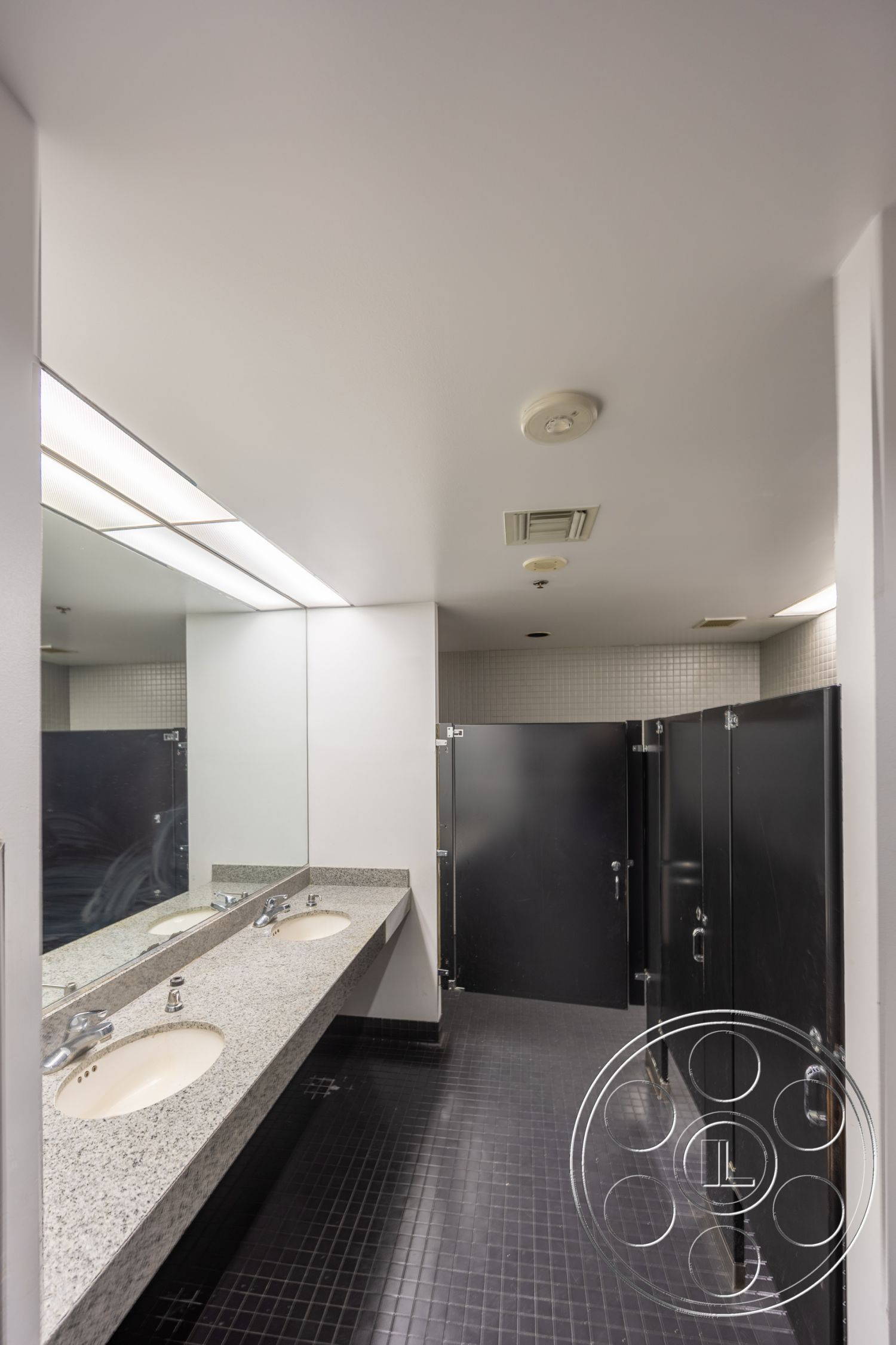 Rooftop 53 - fluorescent lighting, white ceiling, black tile flooring, wall-mounted faucets, interior restroom, granite countertop sinks, mirror above sinks, black stall doors, tiled wall panels
