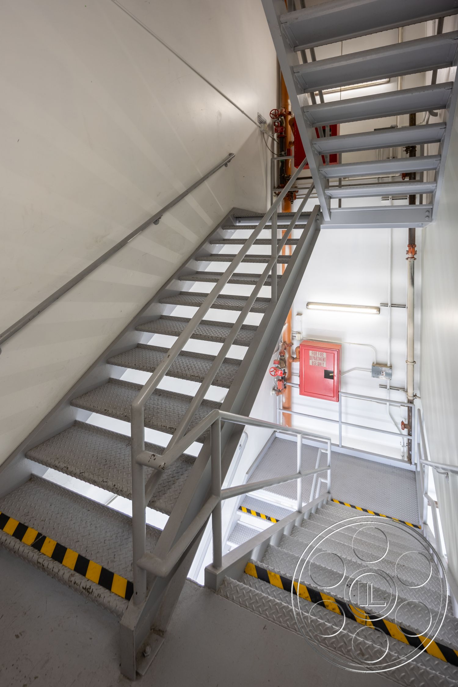 Rooftop 53 - fluorescent lighting, exposed concrete walls, industrial design elements, interior staircase, metal stair treads, steel handrail, diamond plate steel stairs, factory-style stairwell, fire extinguisher equipment, safety yellow and black striped markings