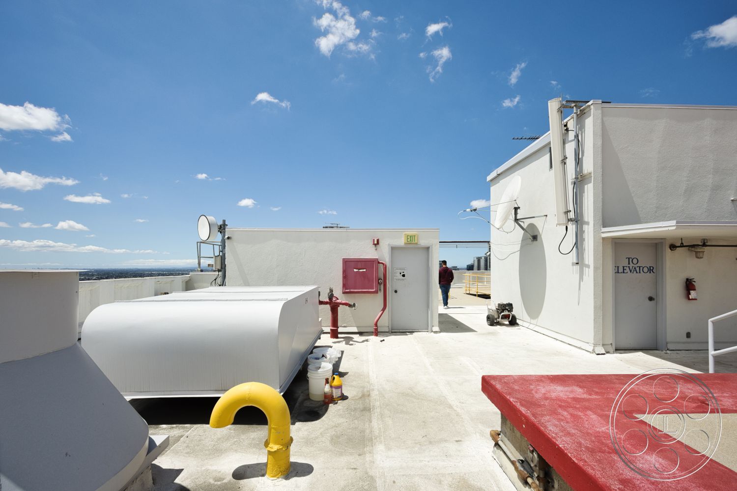 Rooftop 53 - exterior, city skyline view, clear blue sky, white painted walls, industrial rooftop setting, metal piping details, flat concrete rooftop, red fire extinguisher station, elevator access door, satellite dish installation, yellow utility pipe fittings