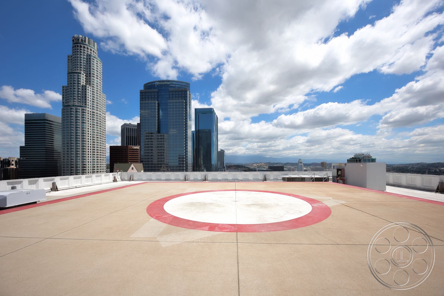 Rooftop 53 - exterior, city view, high-rise buildings, concrete rooftop, modern urban skyline, cloudy blue sky, helicopter landing pad, circular red and white painted helipad, glass skyscraper reflections, metal railing perimeter