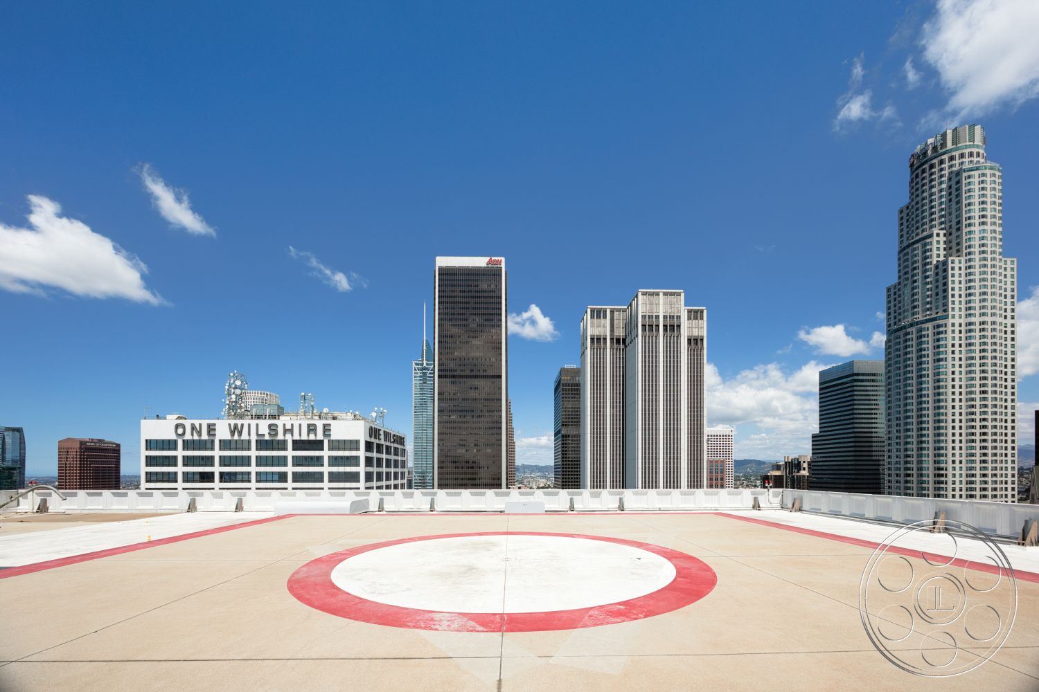 Rooftop 53 - exterior, urban setting, clear blue sky, flat roof design, concrete helipad surface, glass facade buildings, modern skyscraper architecture, red circular helipad marking, skyline view of downtown buildings, clouds in the sky