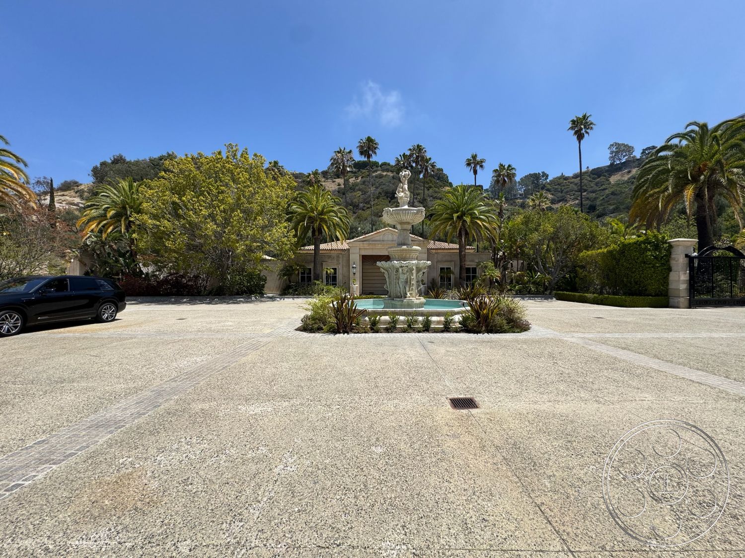 Beverly Compound - exterior, single-story home, symmetrical facade, planted shrubs, tile roof, concrete fountain, stone paver driveway, landscaped garden with palm trees, arched front entrance, garden beds with greenery