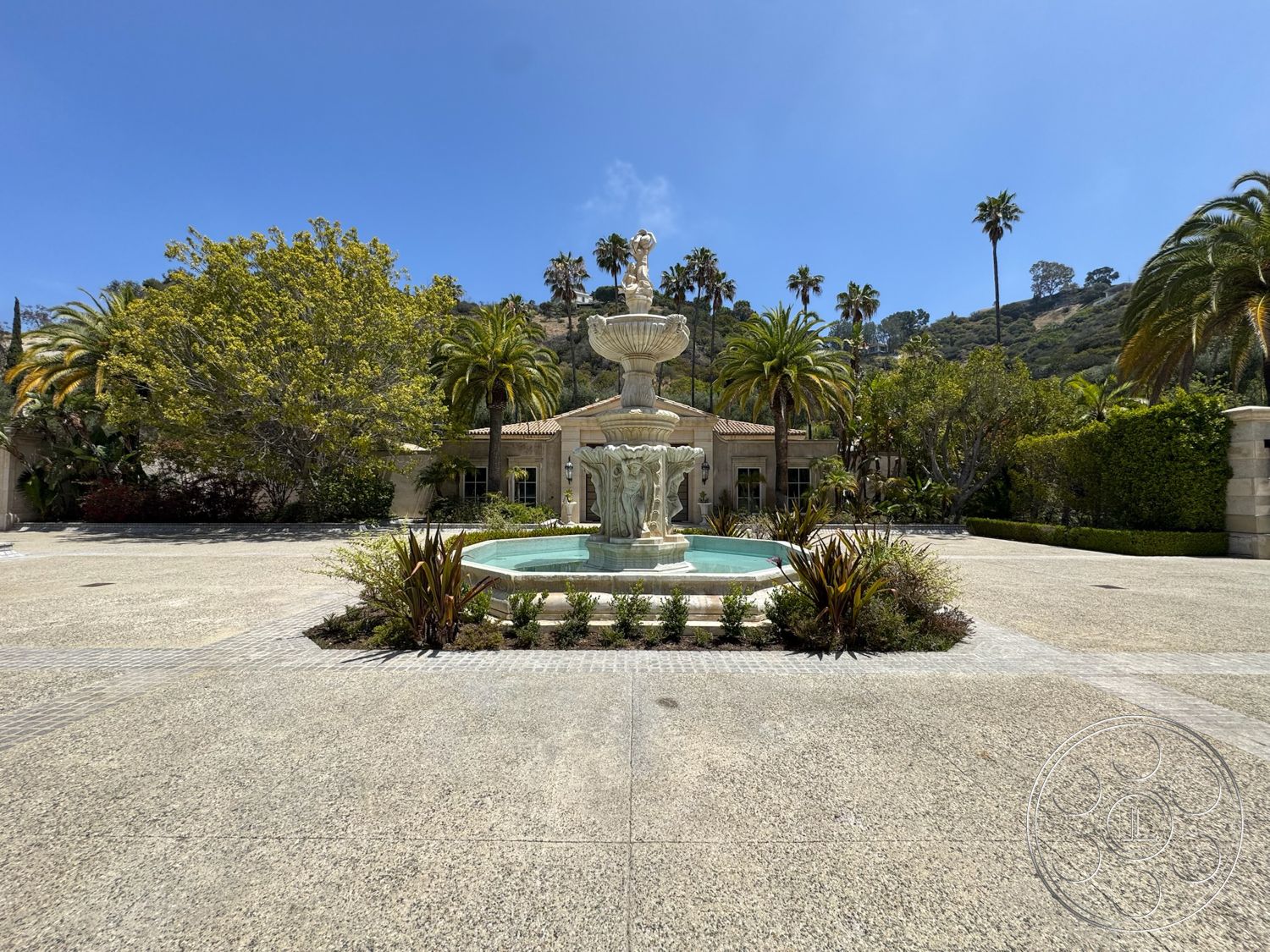 Broad textured forecourt with a tiered stone fountain and tall palms, with a red-tile-roofed range of the estate beyond, at Beverly Compound, a Beverly Hills filming location