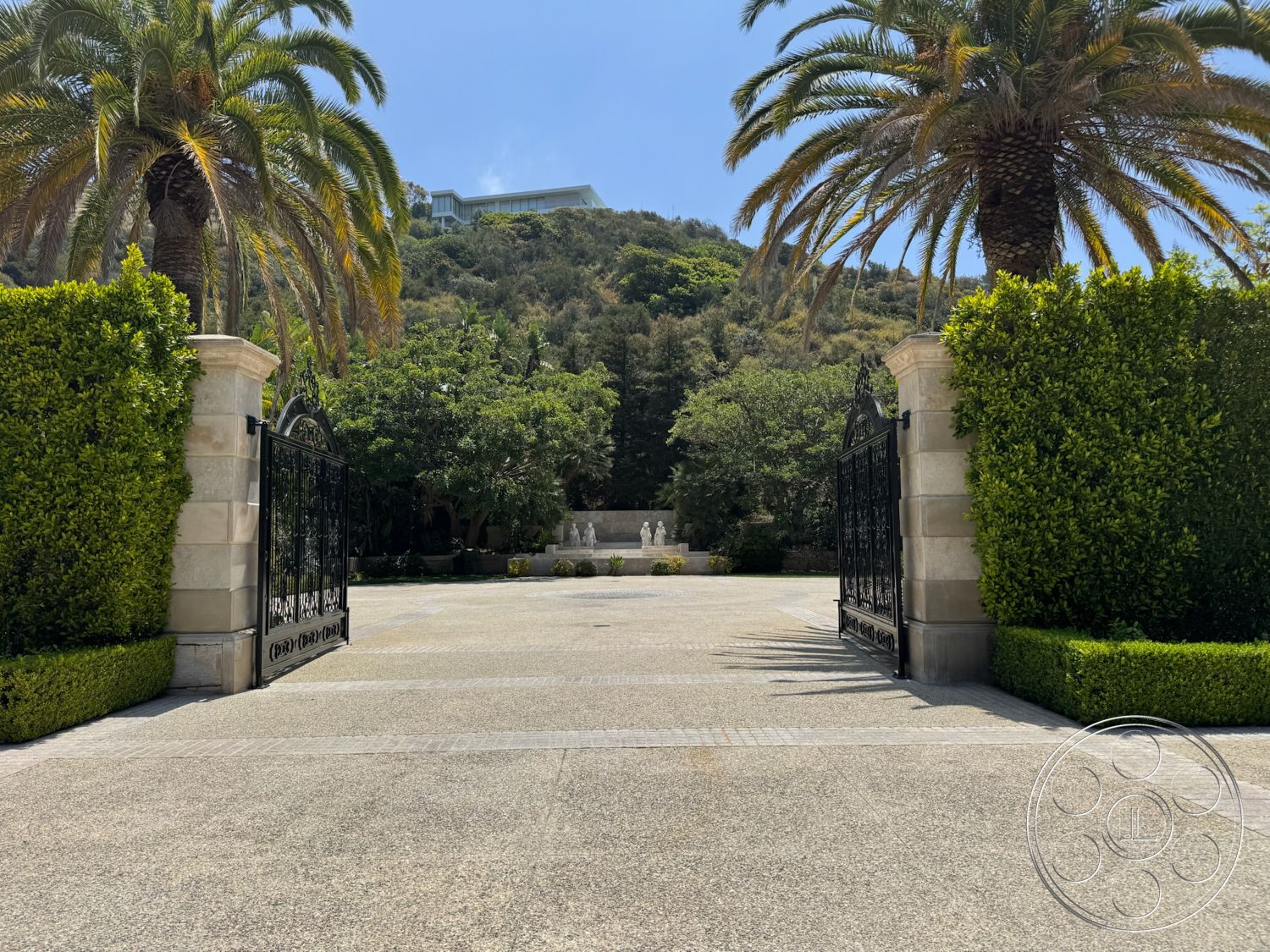 Beverly Compound - exterior, lush greenery, palm trees, landscaped front yard, paved pathway, concrete driveway, fountain feature, boxwood hedges, hillside location, stone gate pillars, decorative wrought iron gate, sunlit entryway