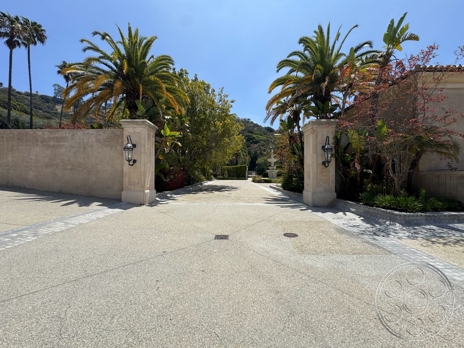 Beverly Compound - exterior, hilltop view, landscaped garden area, exposed aggregate concrete driveway, palms and tropical plants, smooth concrete path, ornamental fountain feature, brick paver border, stucco perimeter wall, arcade lantern light fixtures, entryway with inviting path