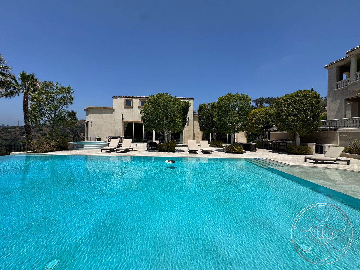 Beverly Compound - exterior, landscaped garden, modern architecture, floor-to-ceiling windows, clear blue sky, infinity pool, tropical palm trees, stone pool deck, sunny outdoor setting, concrete exterior walls, wooden deck chairs, views of surrounding hills