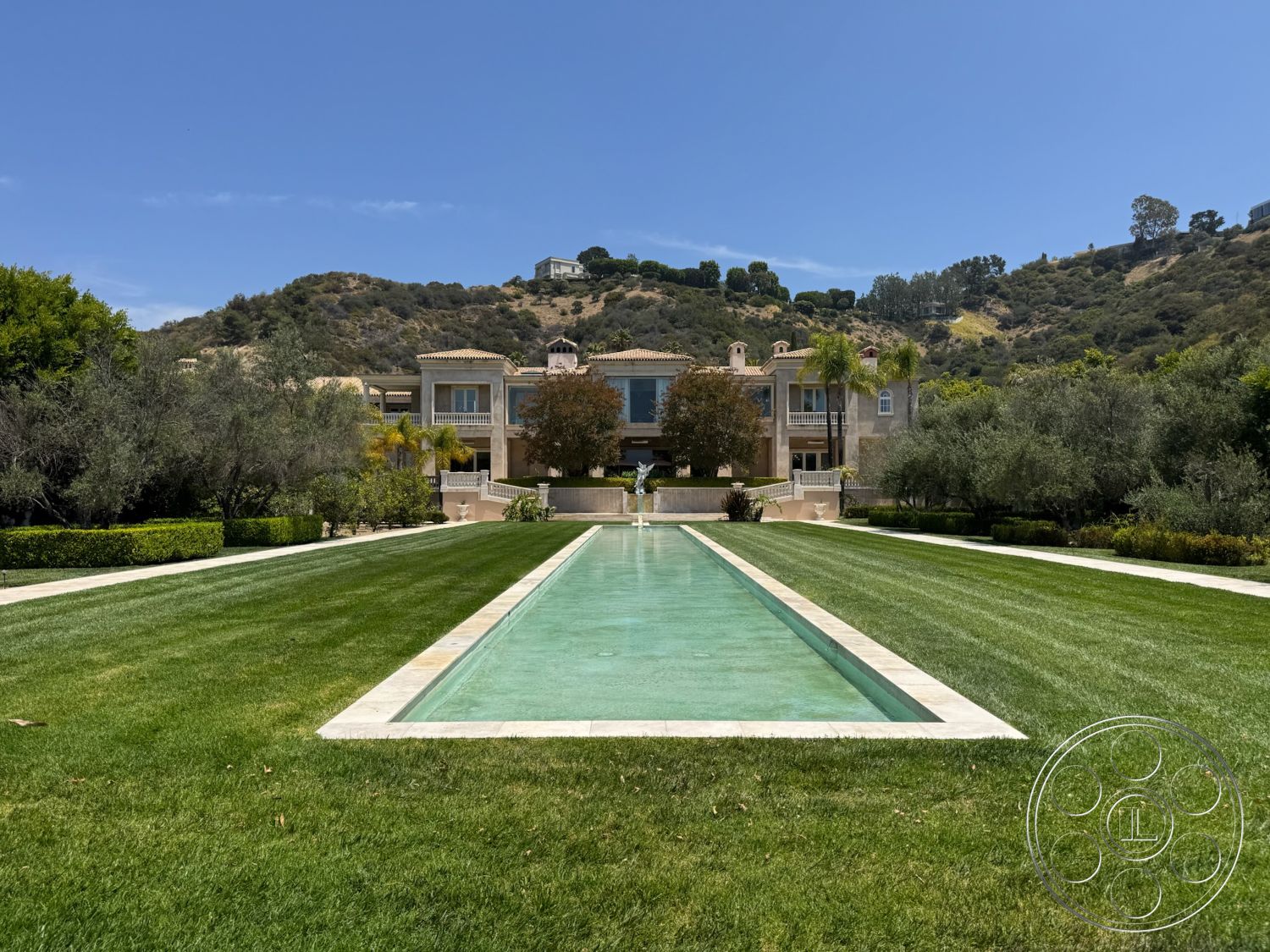 Beverly Compound - exterior, landscaped garden, floor-to-ceiling windows, paved walkway, manicured lawn, stucco facade, symmetrical landscaping, rectangular pool, balcony with wrought iron railings, stone pool deck, planted palm trees, green hillside backdrop, modern Mediterranean home, stone terrace steps, grassy expanse