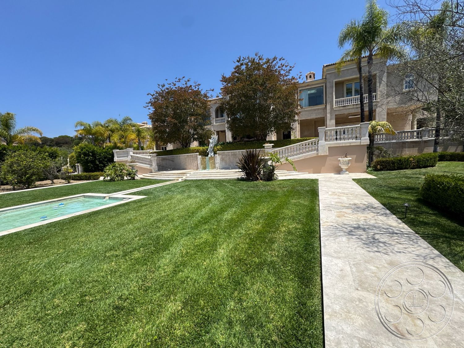 Beverly Compound - exterior, palm trees, rectangular swimming pool, landscaped lawn, clear blue sky, fountain feature, ornamental shrubs, luxurious mansion, formal garden design, terraced garden, steps leading to entrance, sunny outdoor setting, stone exterior walls, marble patio flooring, concrete pool surround, balustrade railing