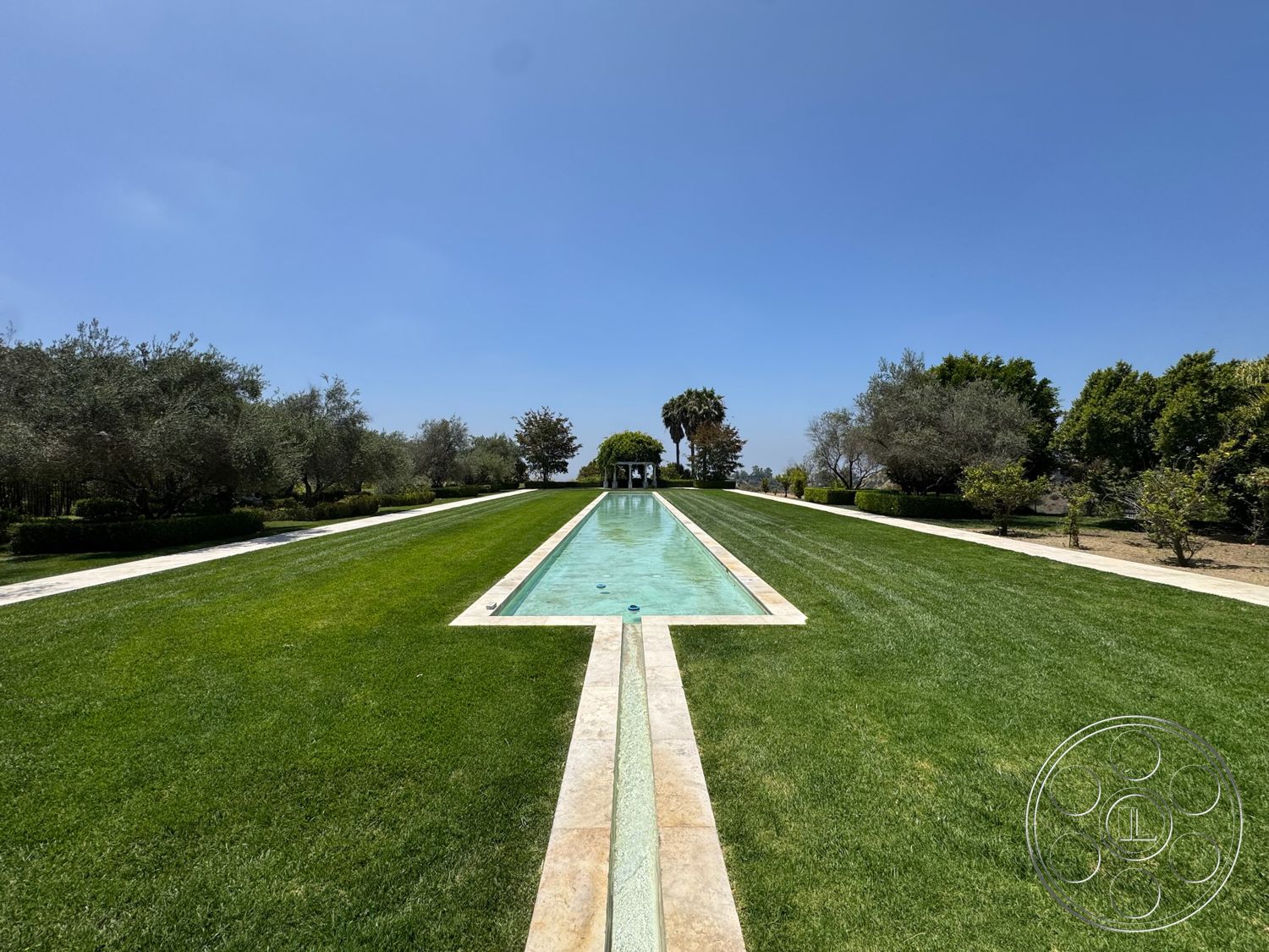 Beverly Compound - exterior, manicured lawn, clear blue sky, symmetrical landscaping, rectangular pool, sunny outdoor setting, horizontally aligned hedges, stone pool border, modern outdoor architecture, tree-lined walkway, view of surrounding greenery