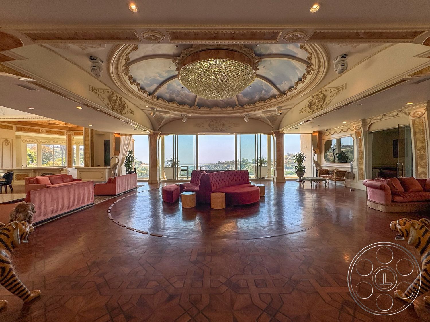 Beverly Compound - view of outdoor landscape, large floor-to-ceiling windows, stylish area rug, curved seating arrangement, ornate wall moldings, floor-to-ceiling mirrors, open plan living space, decorative coffered ceiling with chandelier, intricately painted ceiling with clouds, glossy hardwood flooring, pink velvet sofas, round wooden accent tables, luxurious lounge area, elegant art deco design elements