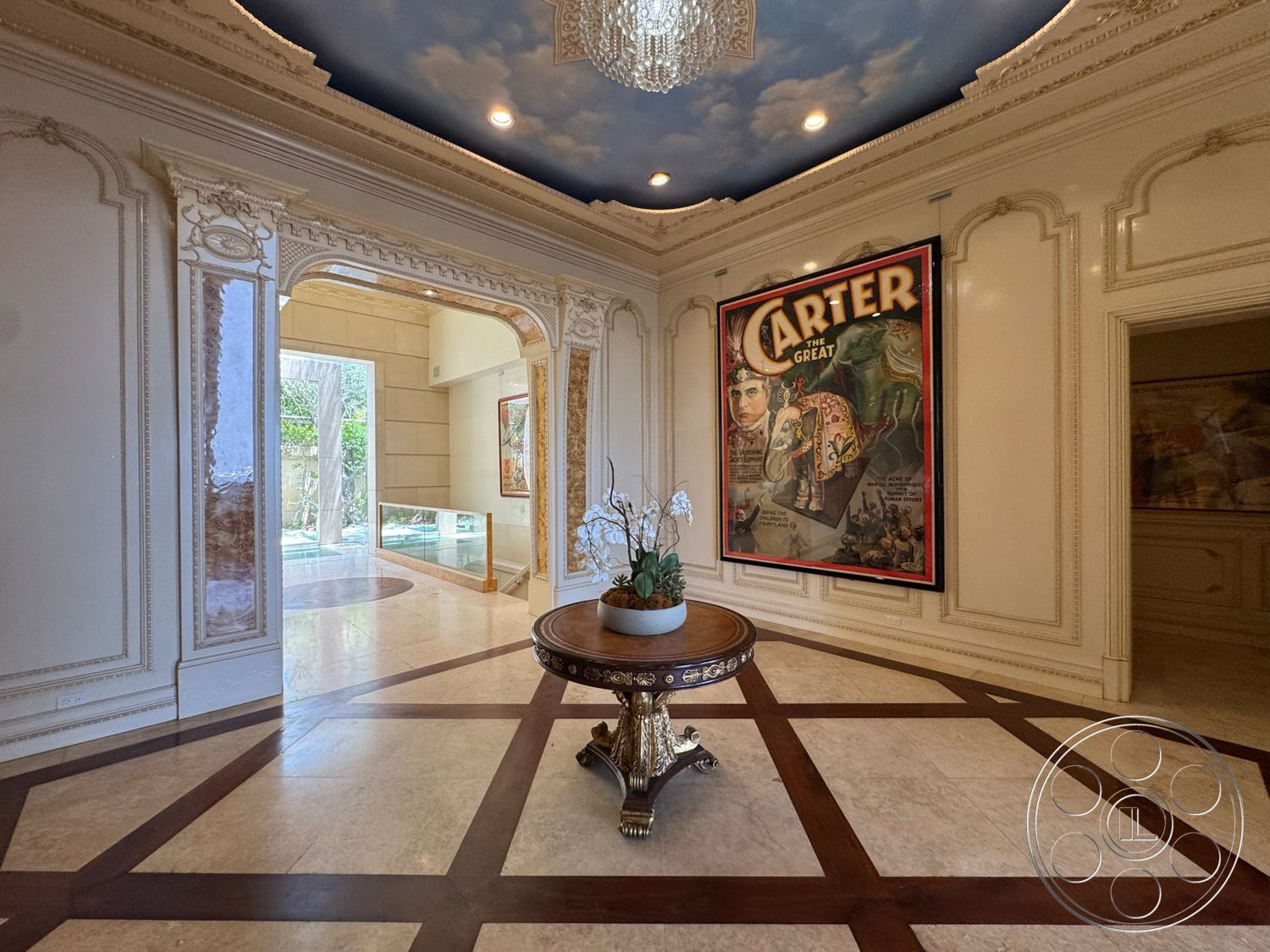 Beverly Compound - glass sliding doors, marble flooring, elegant chandelier, intricate crown molding, exposed ceiling with cloud mural, framed vintage poster art, ornate baseboards, wood accent table with gold details, geometric wood inlay pattern, decorative floral arrangement on table